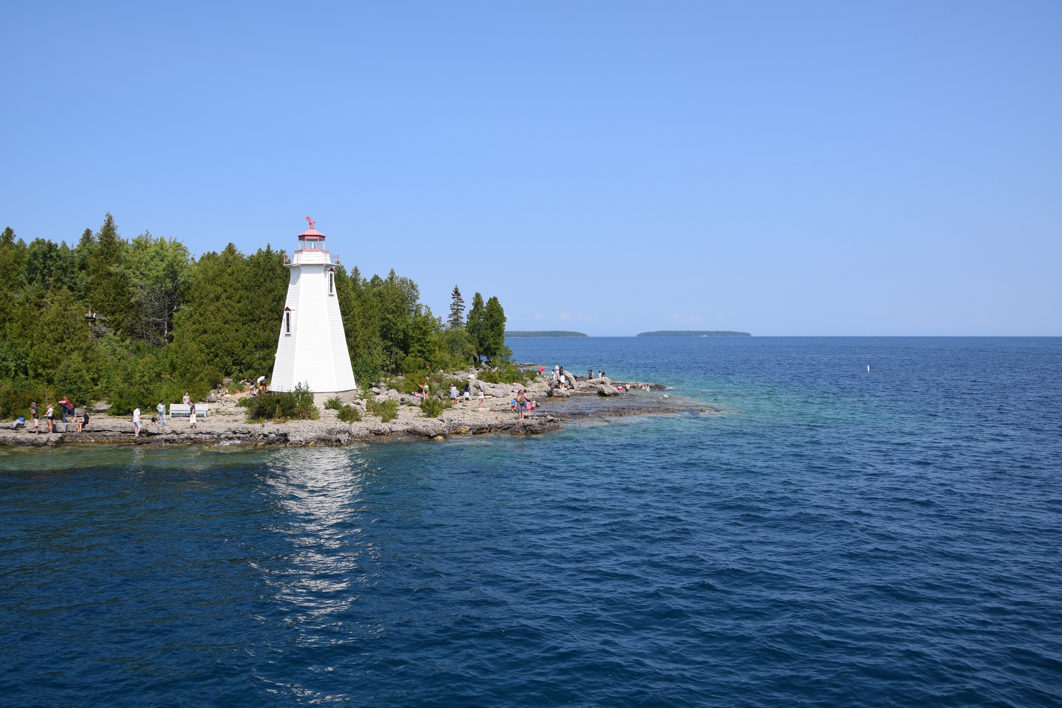 White lighthouse on rocky shore with trees and blue water HD Wallpaper