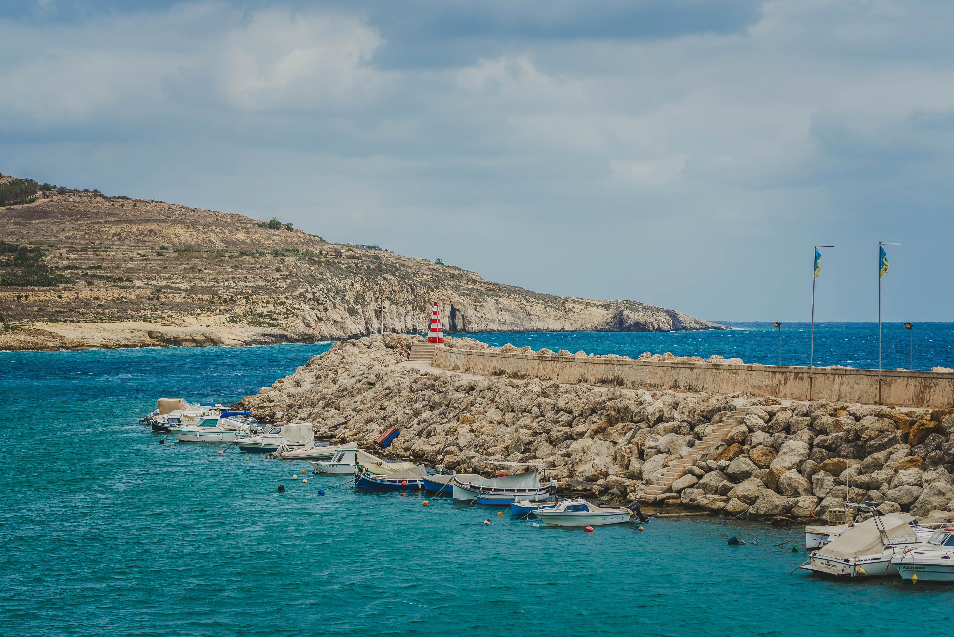 Boats docked at a rocky harbor with a lighthouse on Gozo, Malta HD Wallpaper