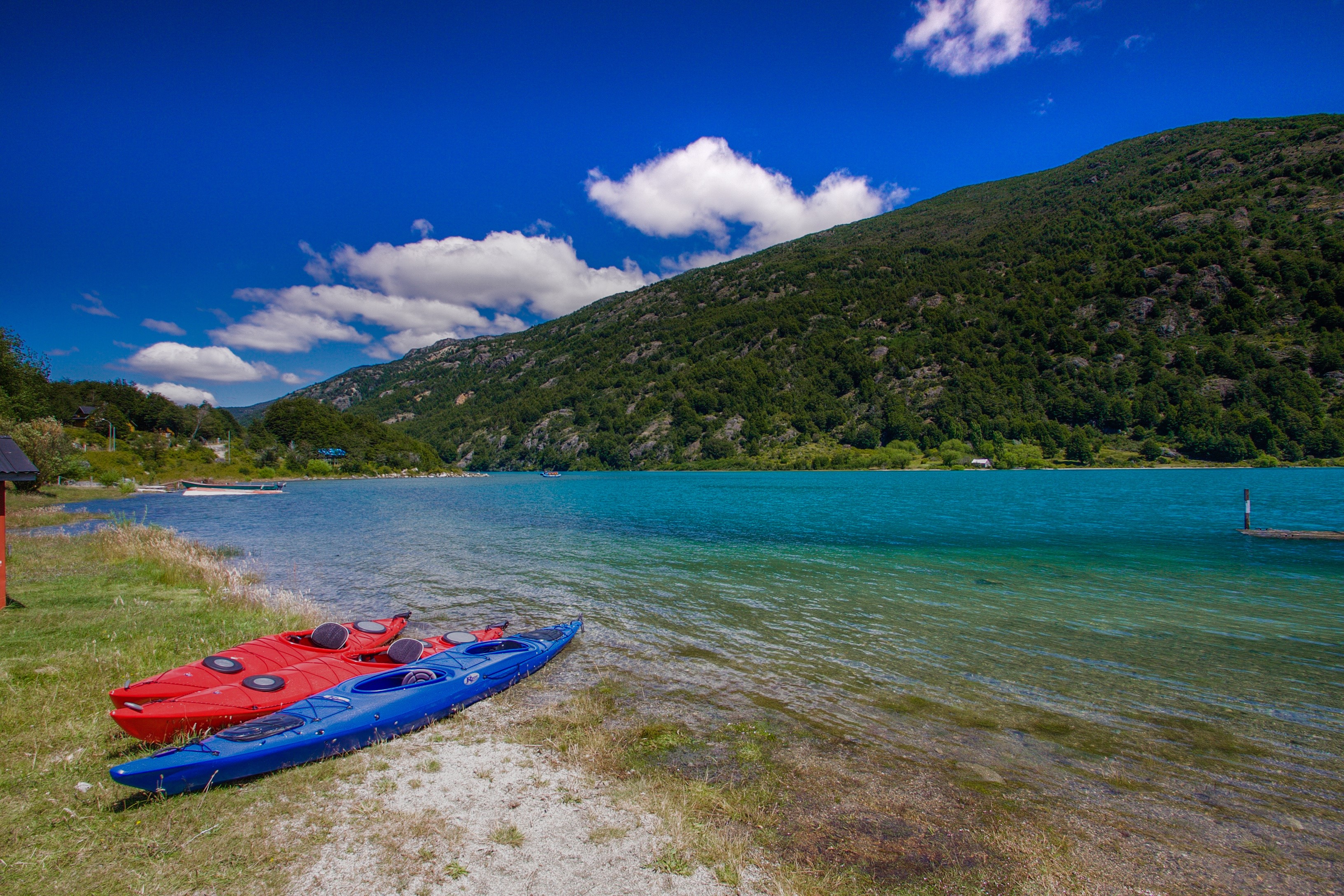 Two kayaks on a sandy shore next to a turquoise lake and forested mountain HD Wallpaper