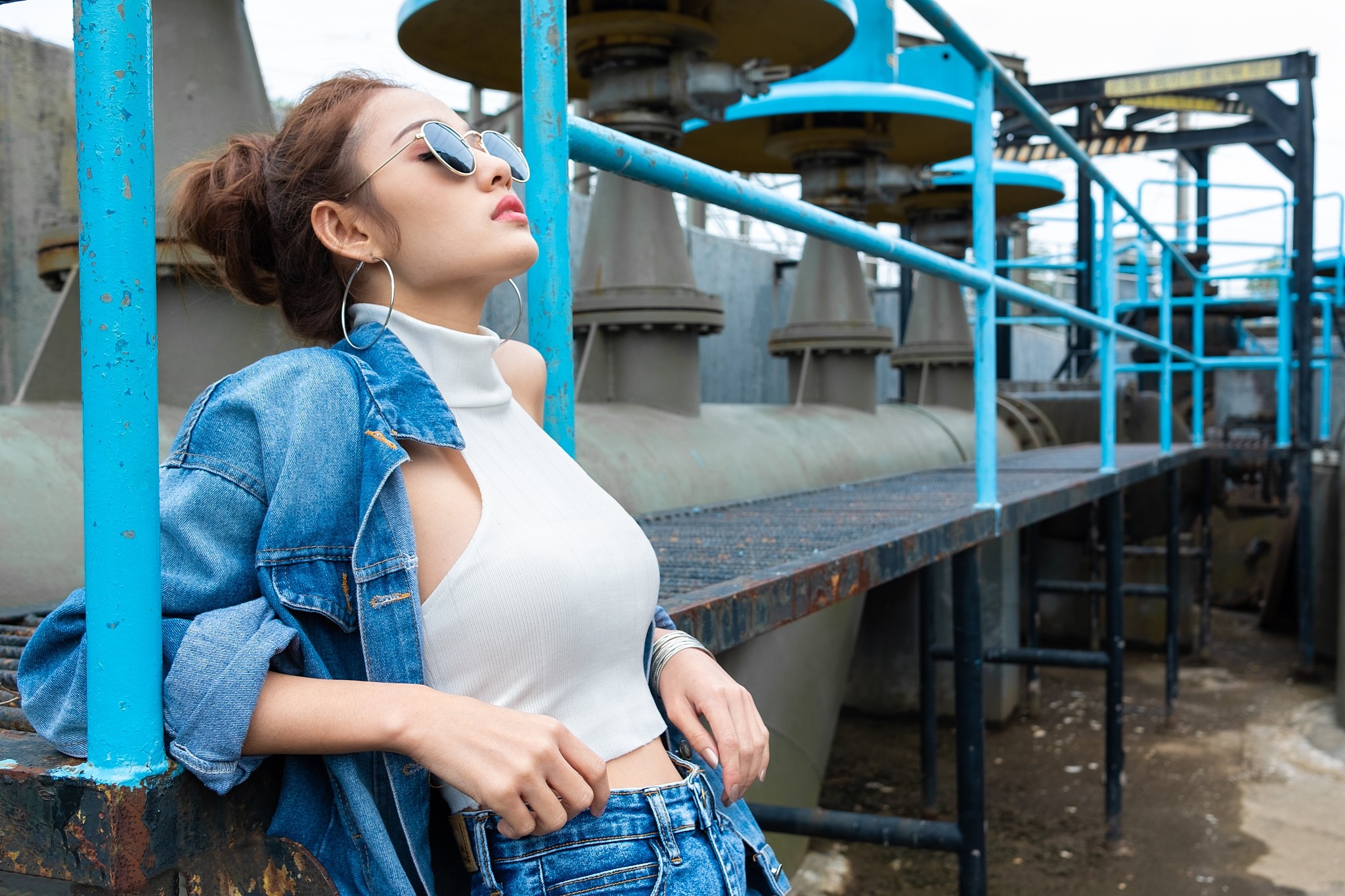 Asian woman in denim jacket and sunglasses posing near industrial pipes HD Wallpaper