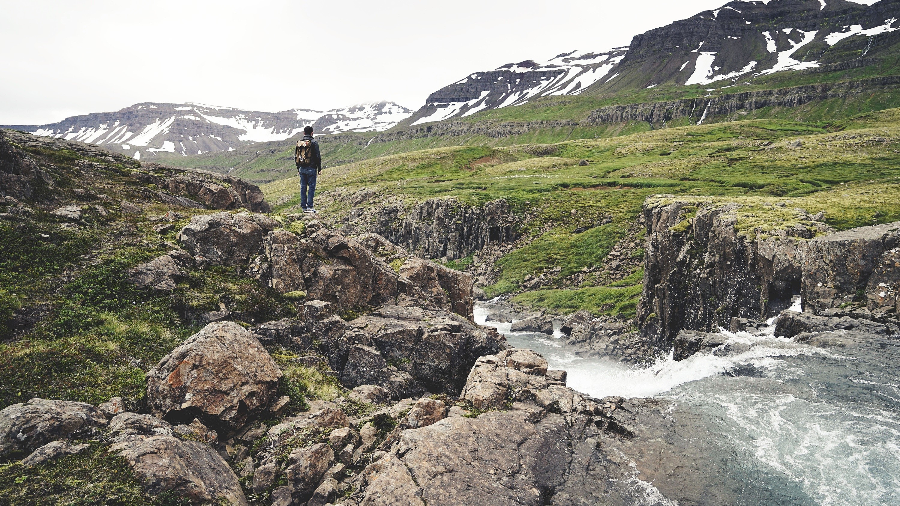 Man with backpack on rocky terrain overlooking mountains and stream HD Wallpaper