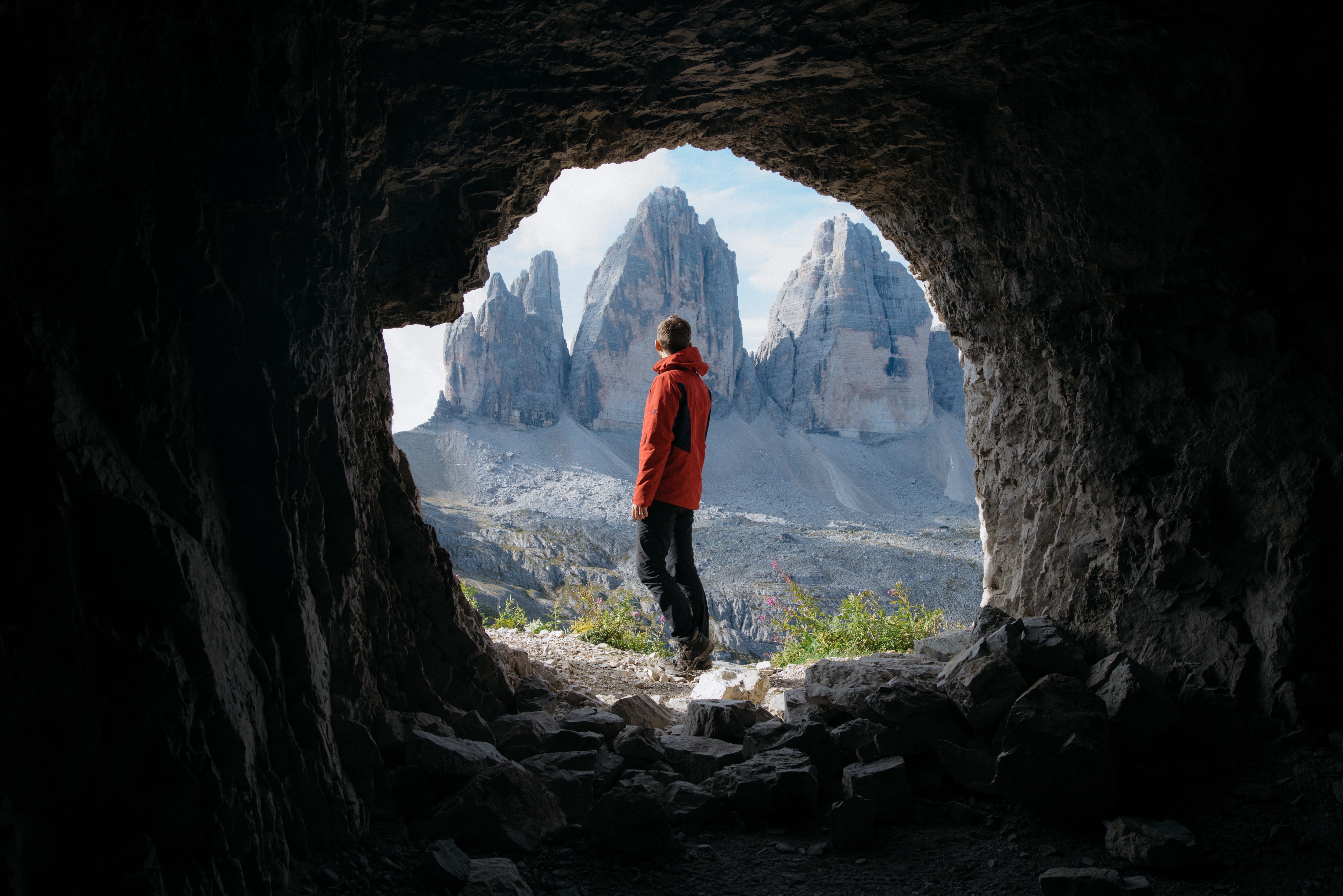 Man in red jacket looking at mountains from cave HD Wallpaper