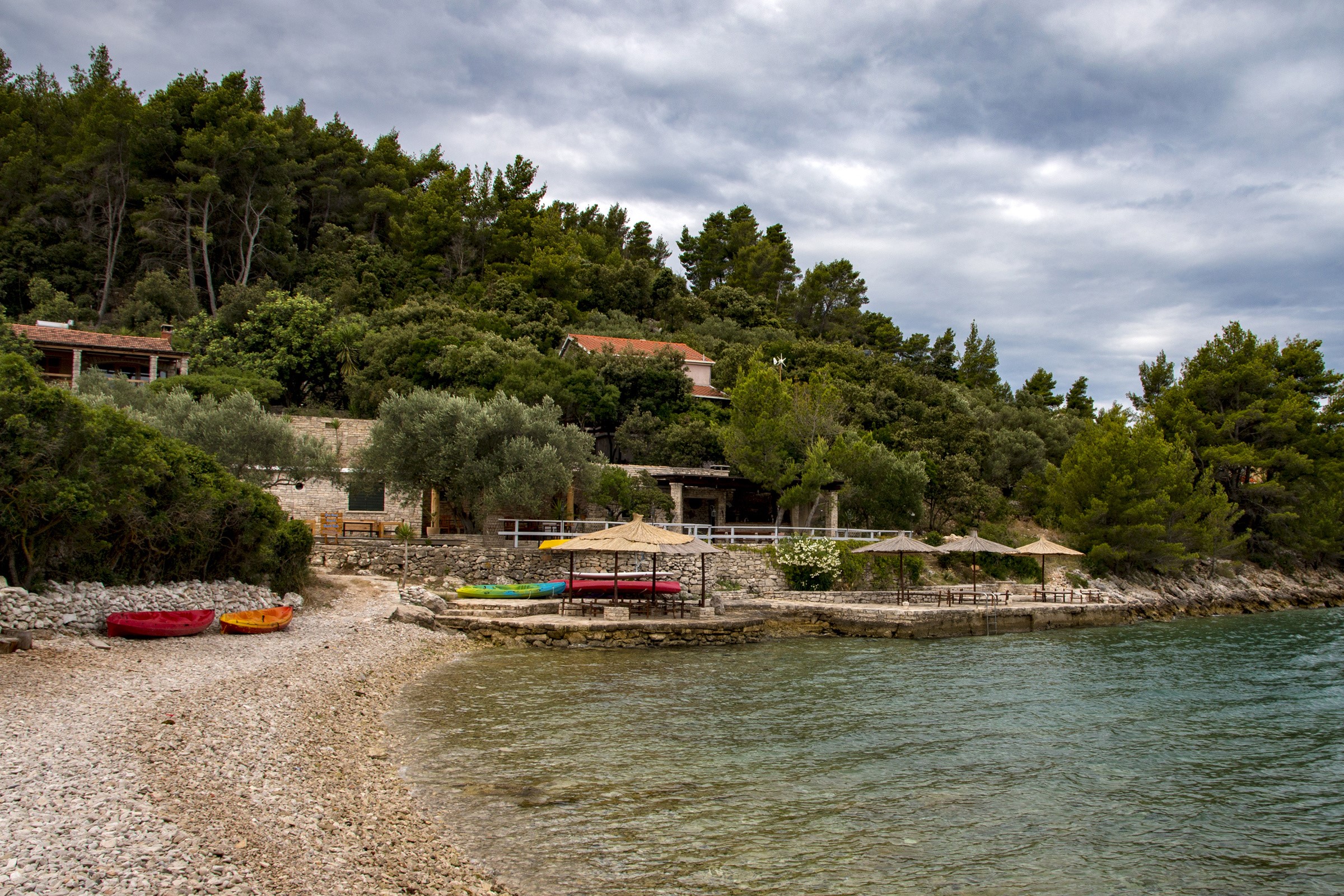 Kayaks on a pebble beach with trees and buildings in Croatia HD Wallpaper