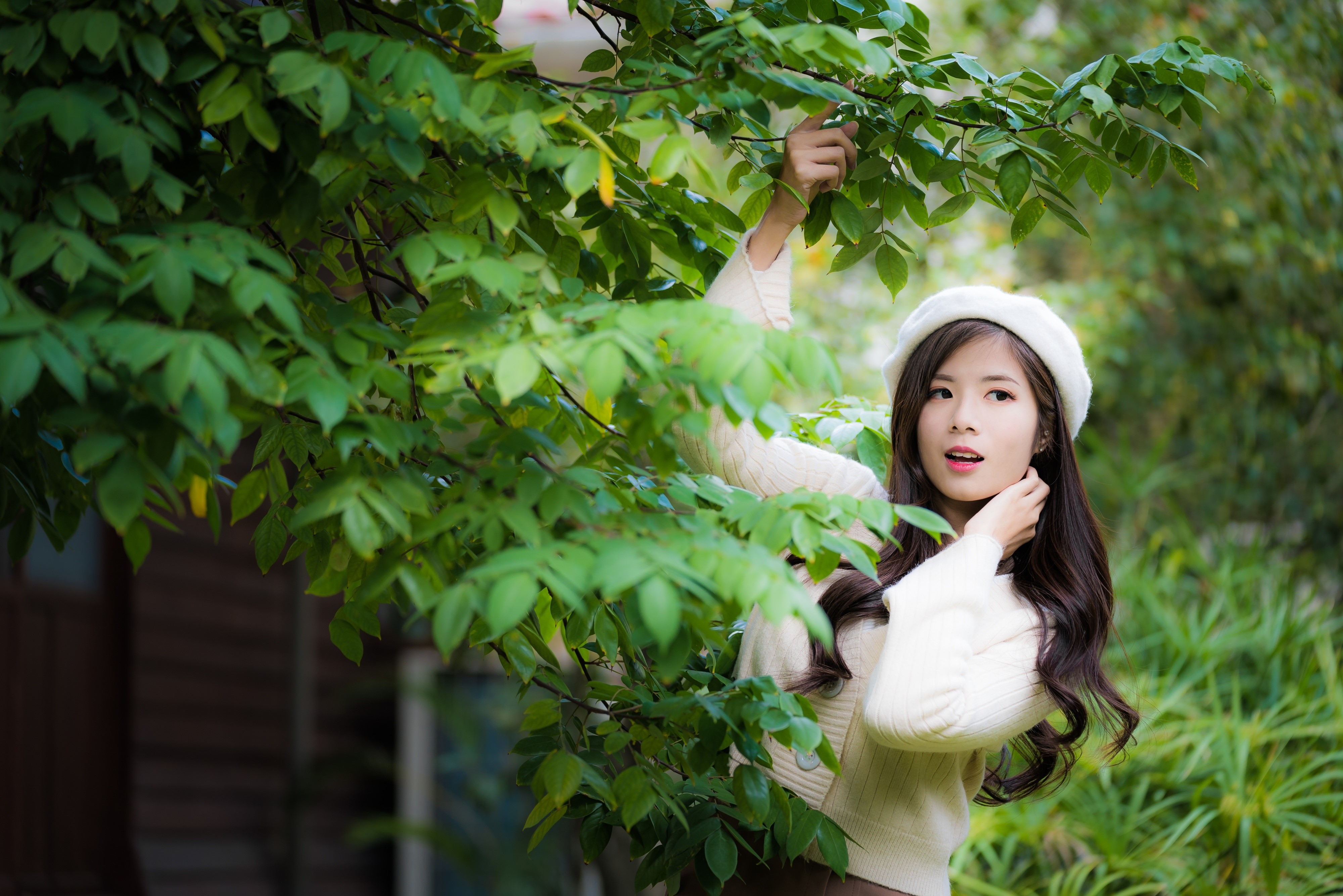 Asian woman in sweater and beret behind foliage HD Wallpaper