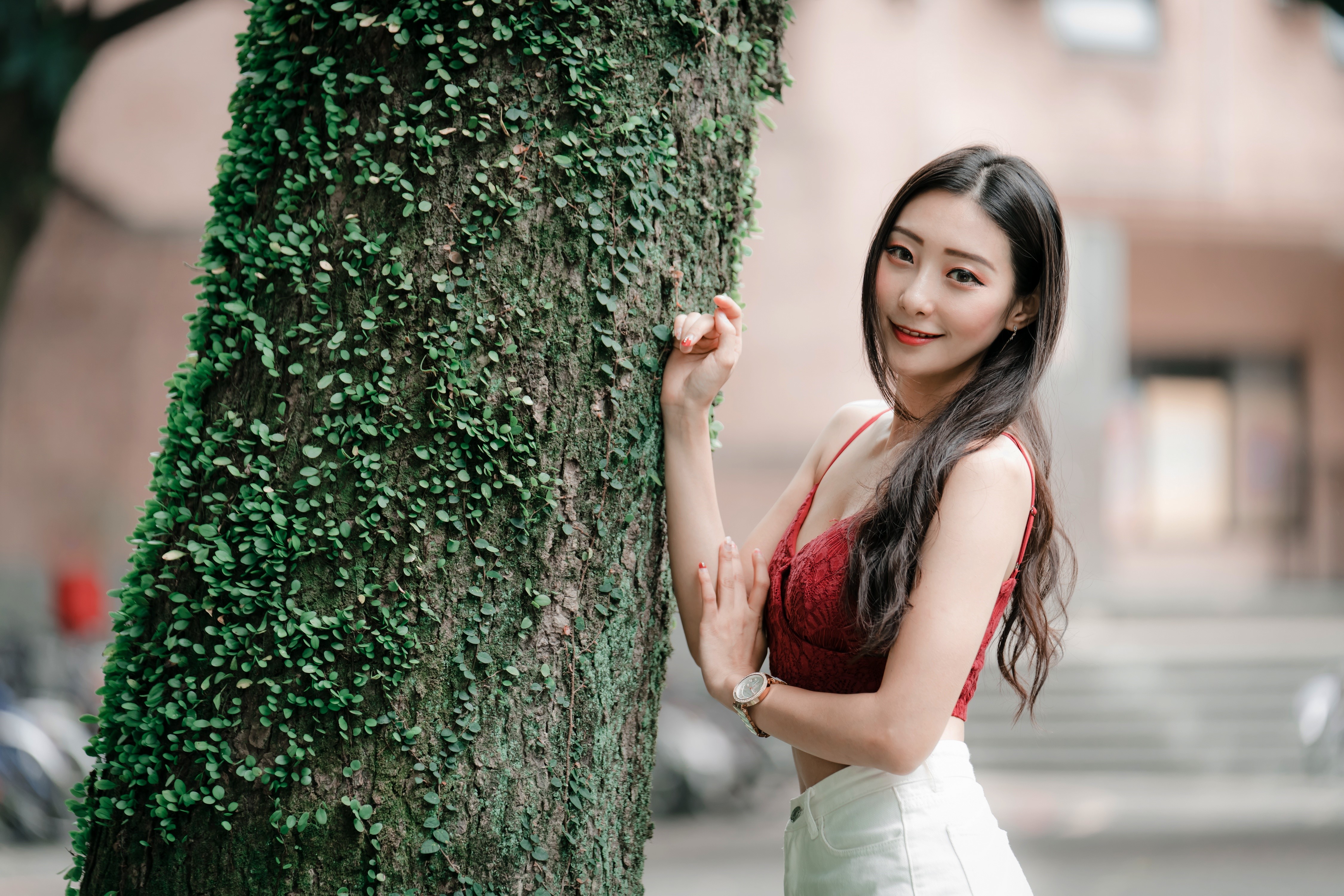 Asian woman in red top and white shorts by tree trunk HD Wallpaper
