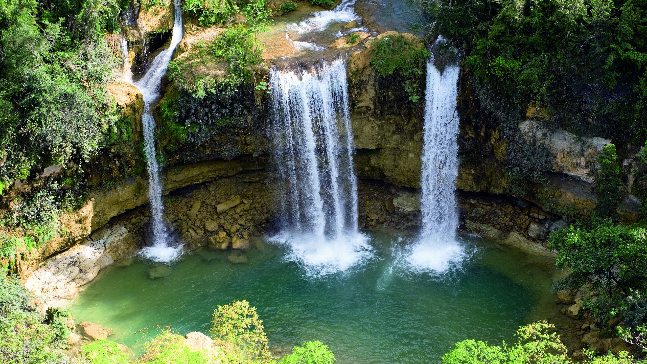 Multiple waterfalls cascade into a turquoise pool surrounded by greenery HD Wallpaper