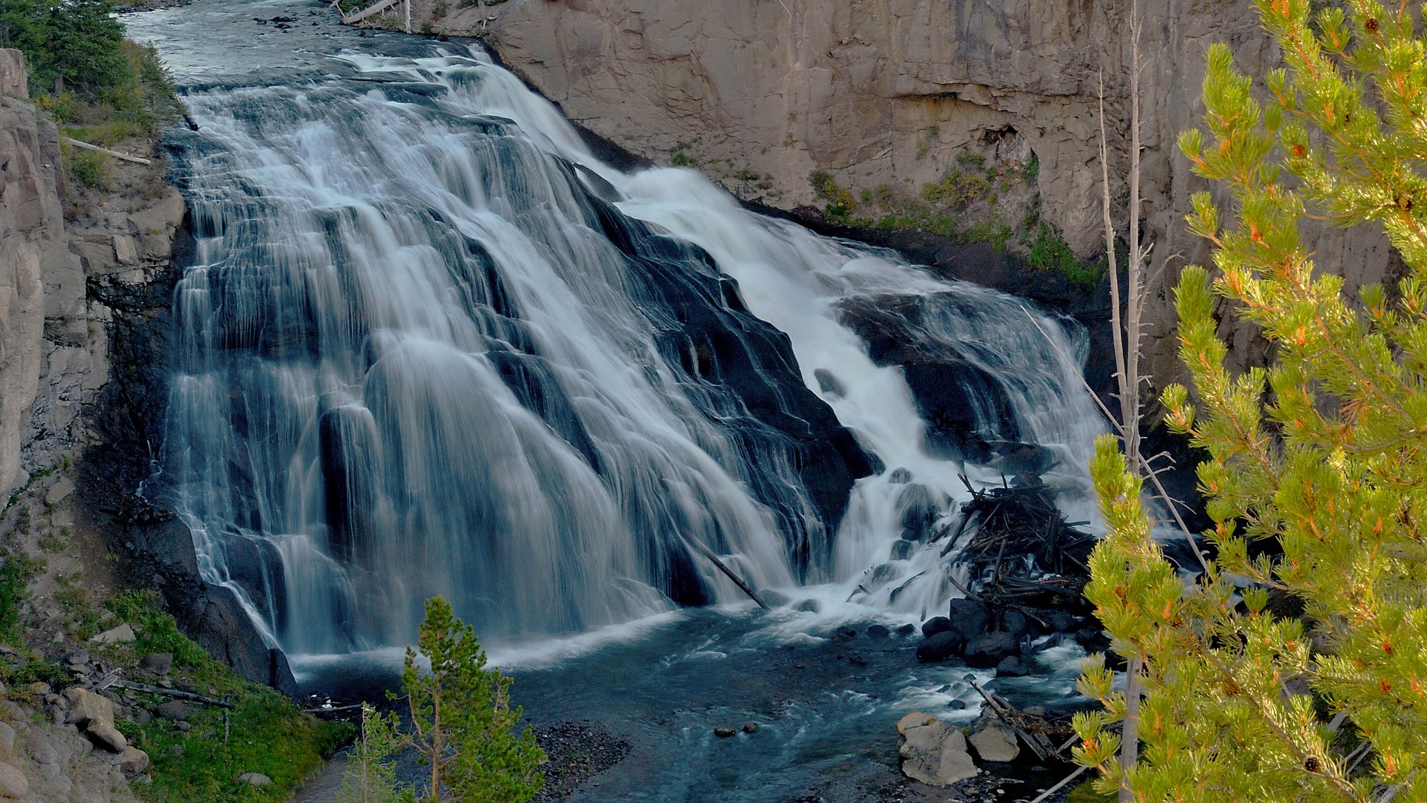Waterfall cascading over dark rocks with pine trees HD Wallpaper