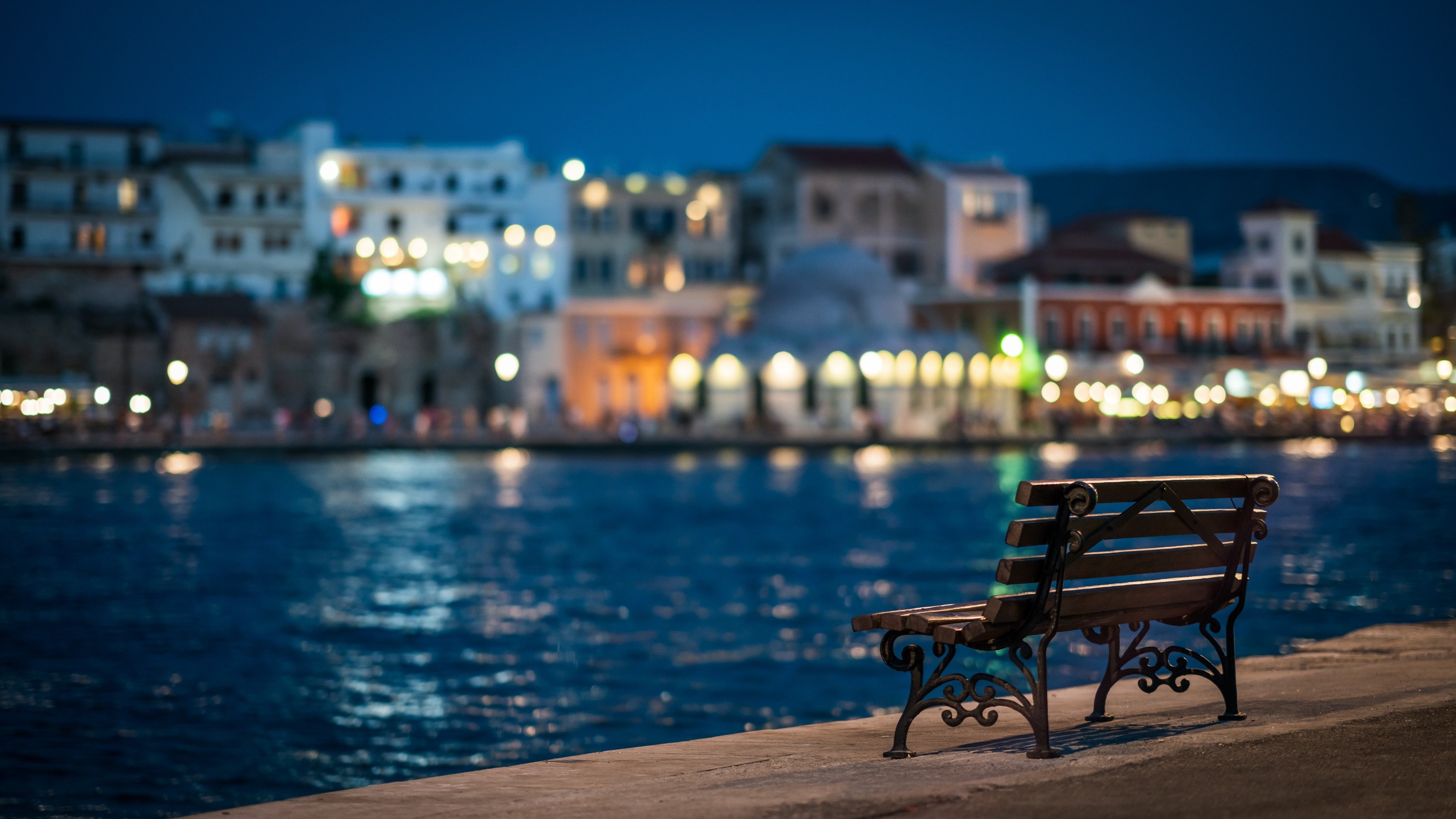 Bench by the water at night with bokeh lights HD Wallpaper