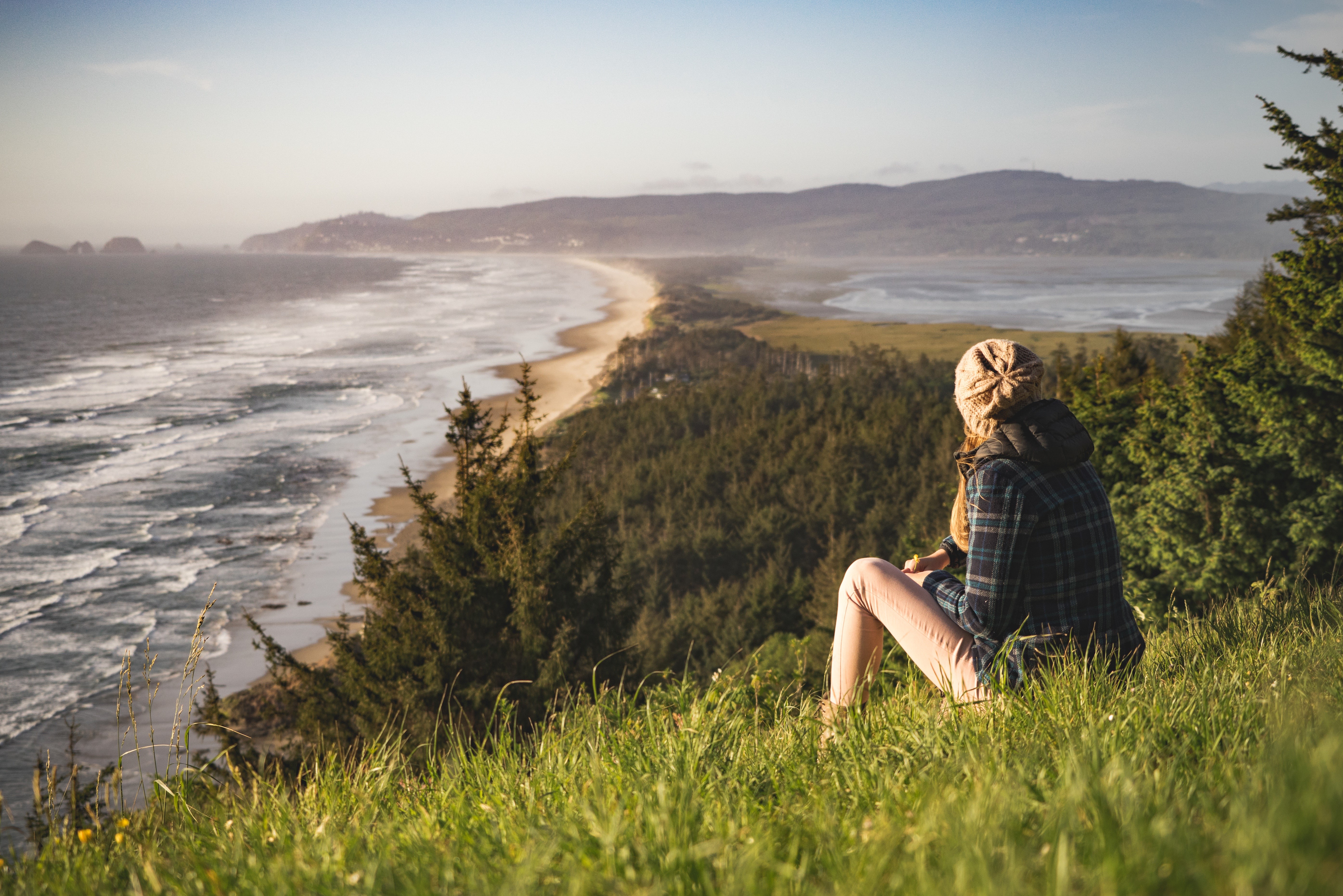 Woman in winter hat sitting on grass overlooking sea coast HD Wallpaper
