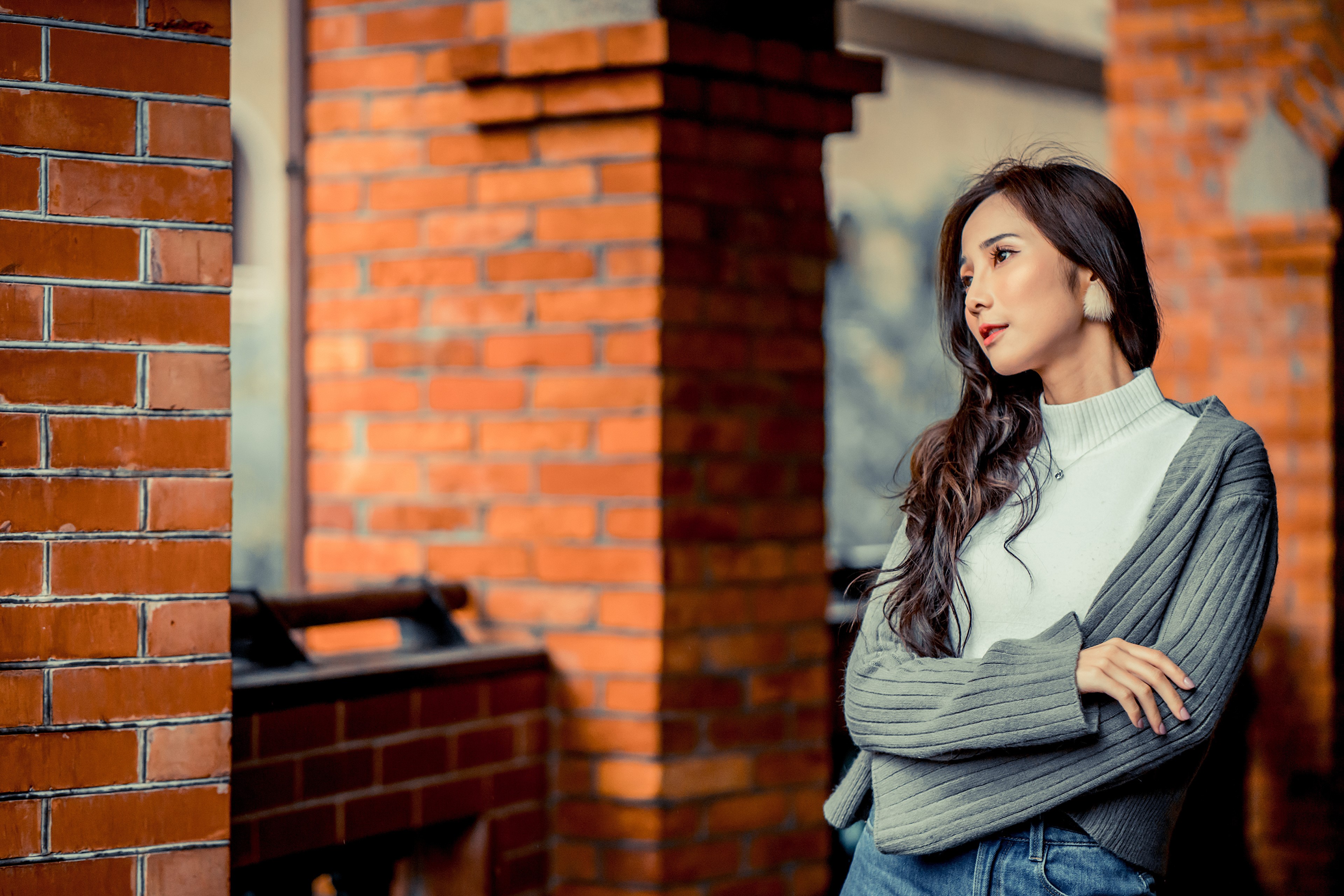 Asian woman in sweater and jeans posing near brick wall HD Wallpaper