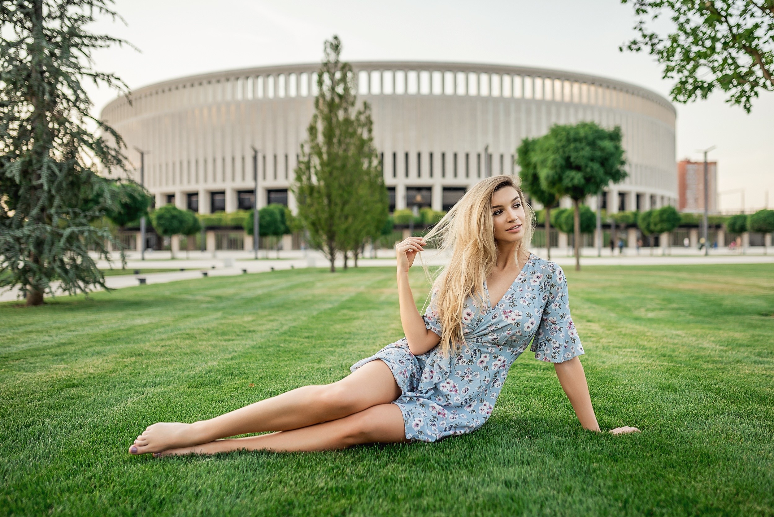 Blonde girl in a dress sitting on grass in front of a stadium HD Wallpaper