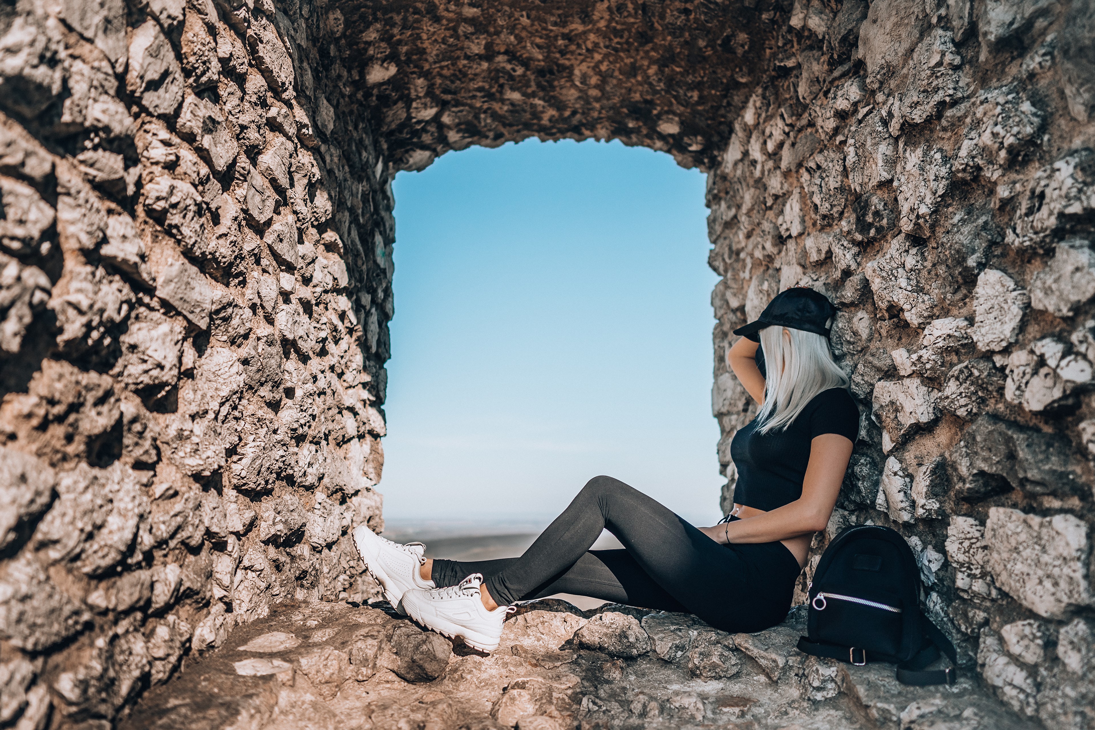 Blonde girl in baseball cap sitting by stone wall HD Wallpaper