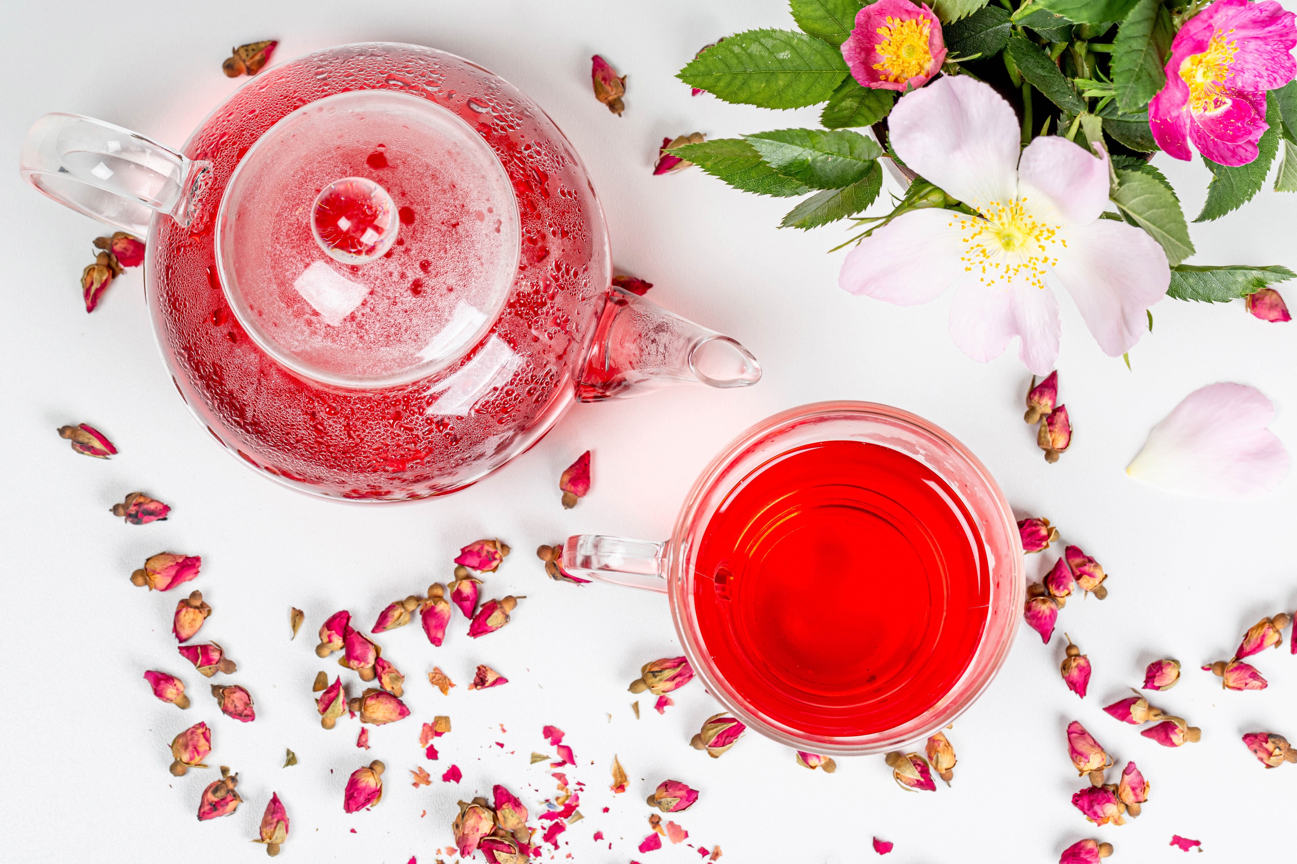 Glass teapot and mug filled with red tea, surrounded by rose buds and flowers HD Wallpaper