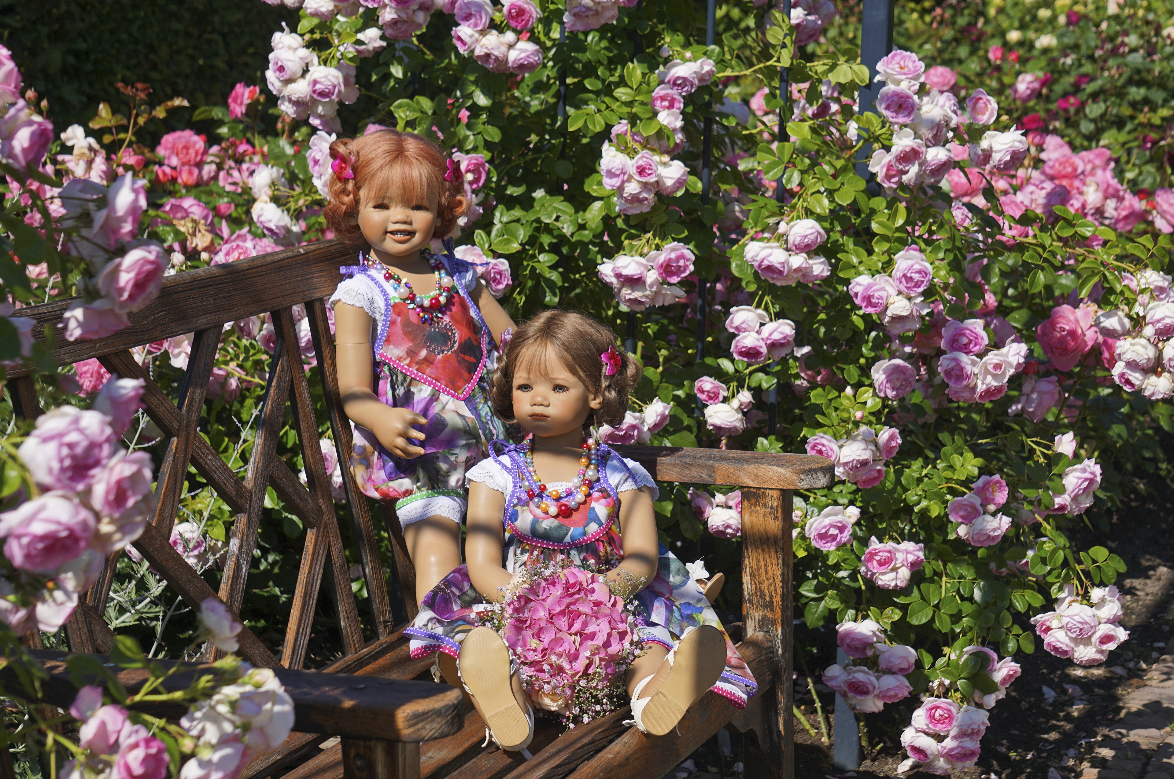 Two dolls sitting on a bench in a rose garden HD Wallpaper