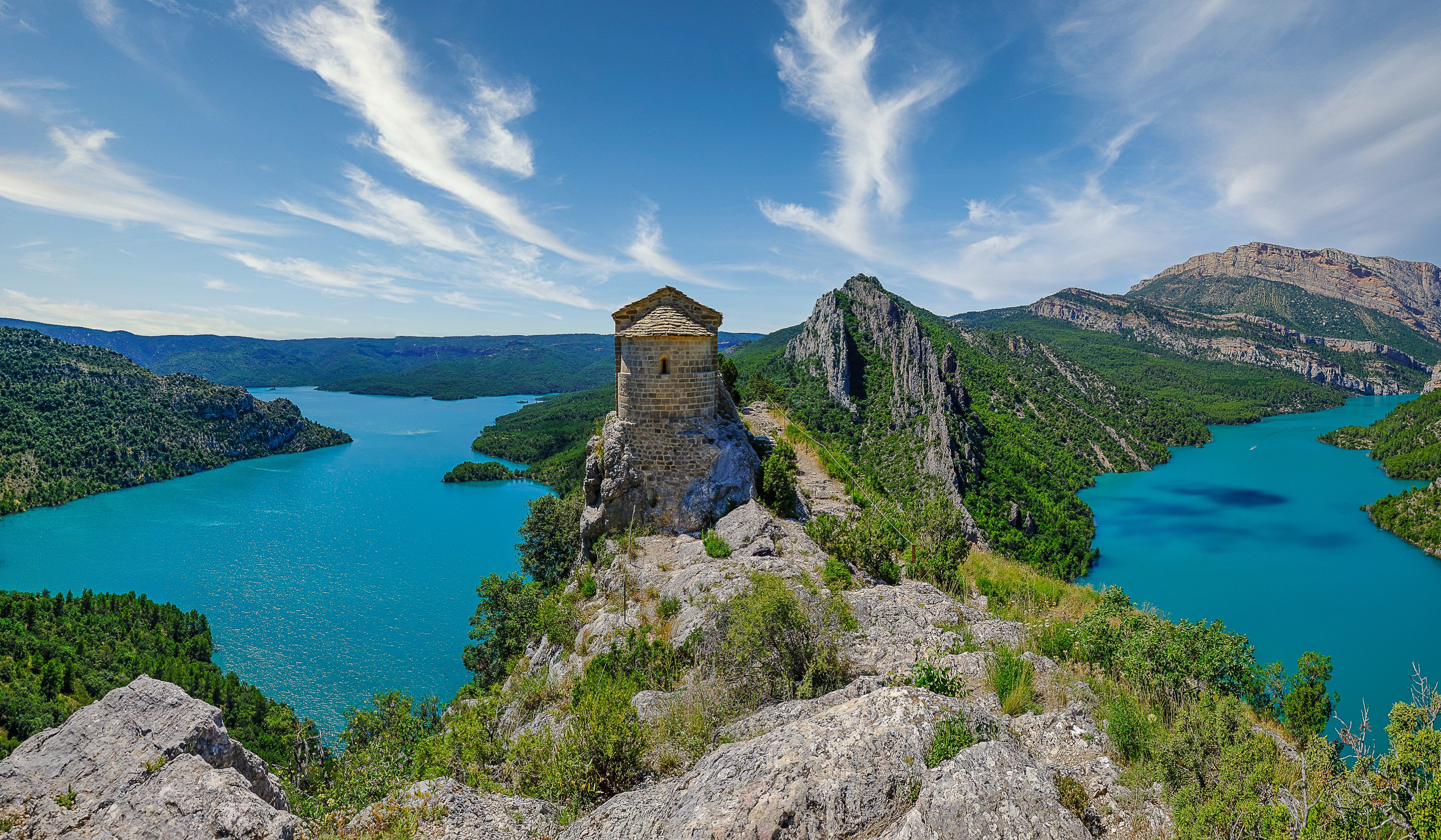 Stone hermitage on a rocky outcrop overlooking a turquoise reservoir in Spain HD Wallpaper