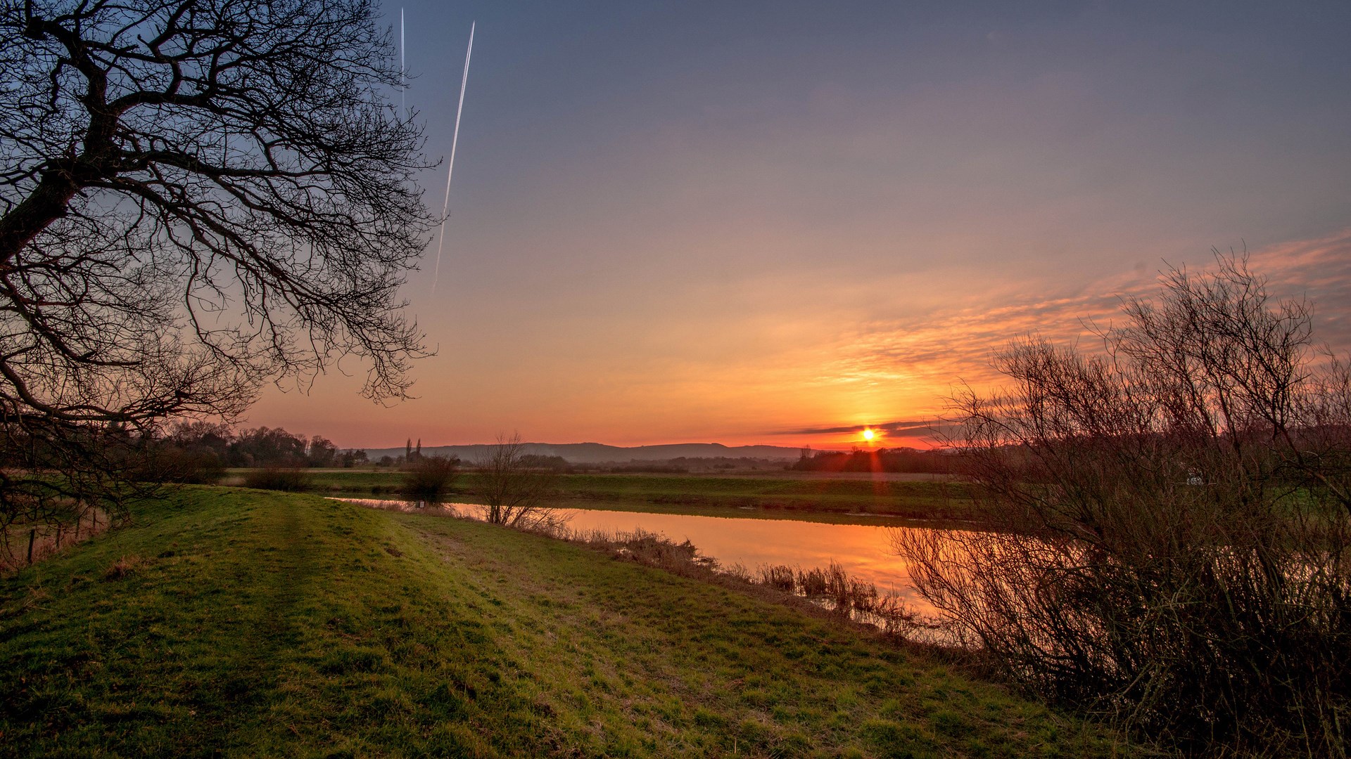 Sunset over a river with trees and contrails HD Wallpaper