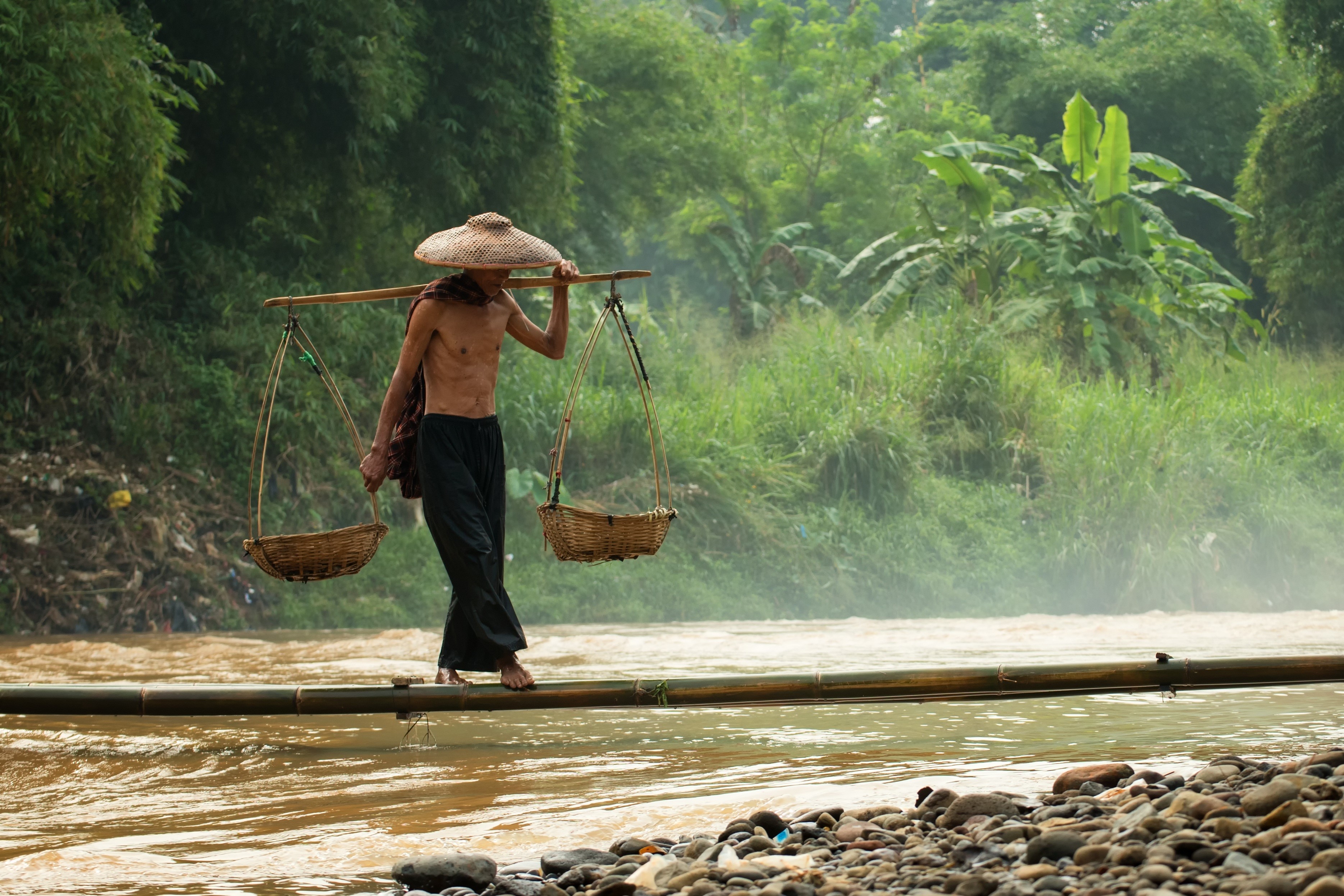 Man carrying baskets across a bamboo bridge over a river HD Wallpaper