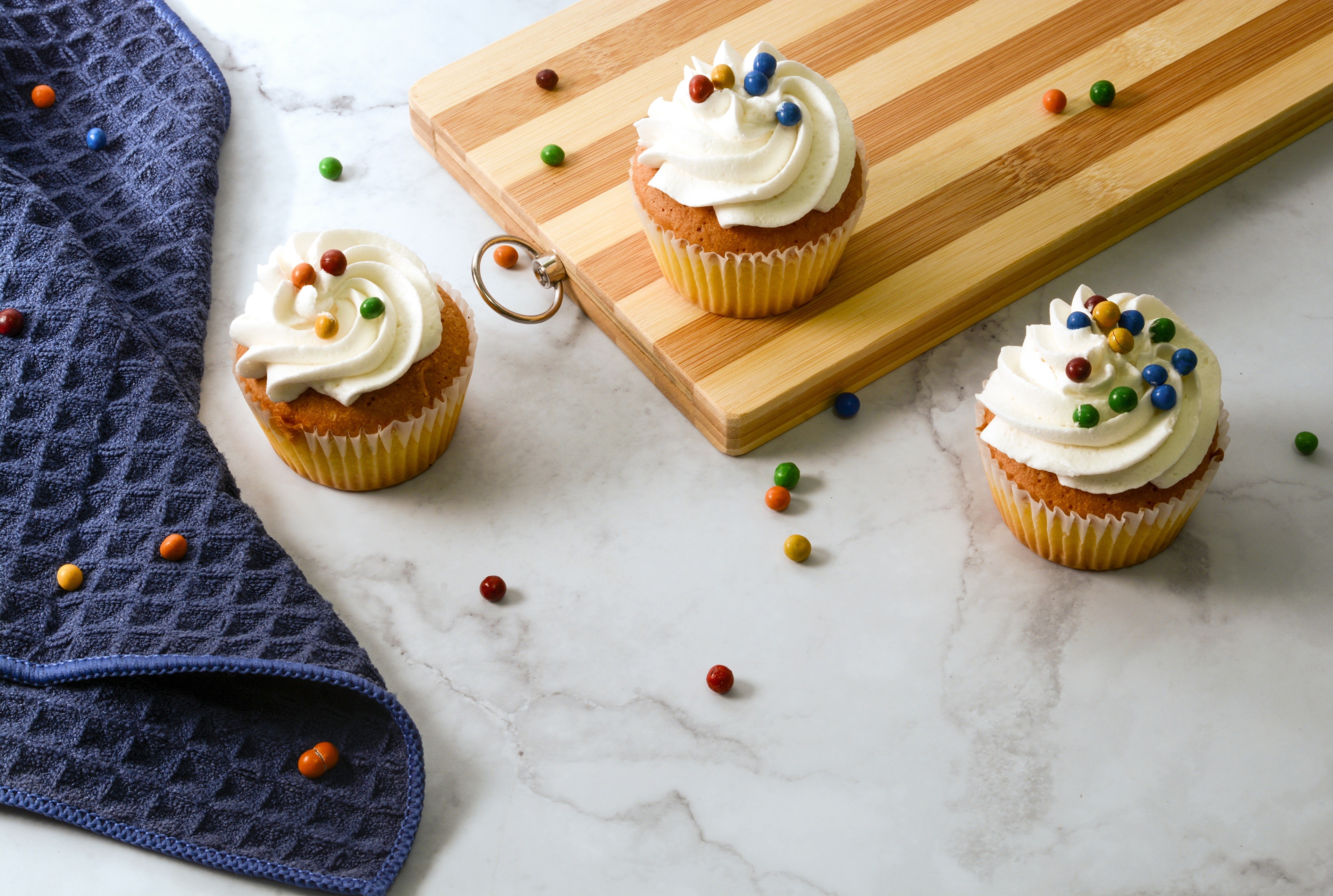 Three cupcakes with white frosting and candy on a marble surface with a cutting board HD Wallpaper