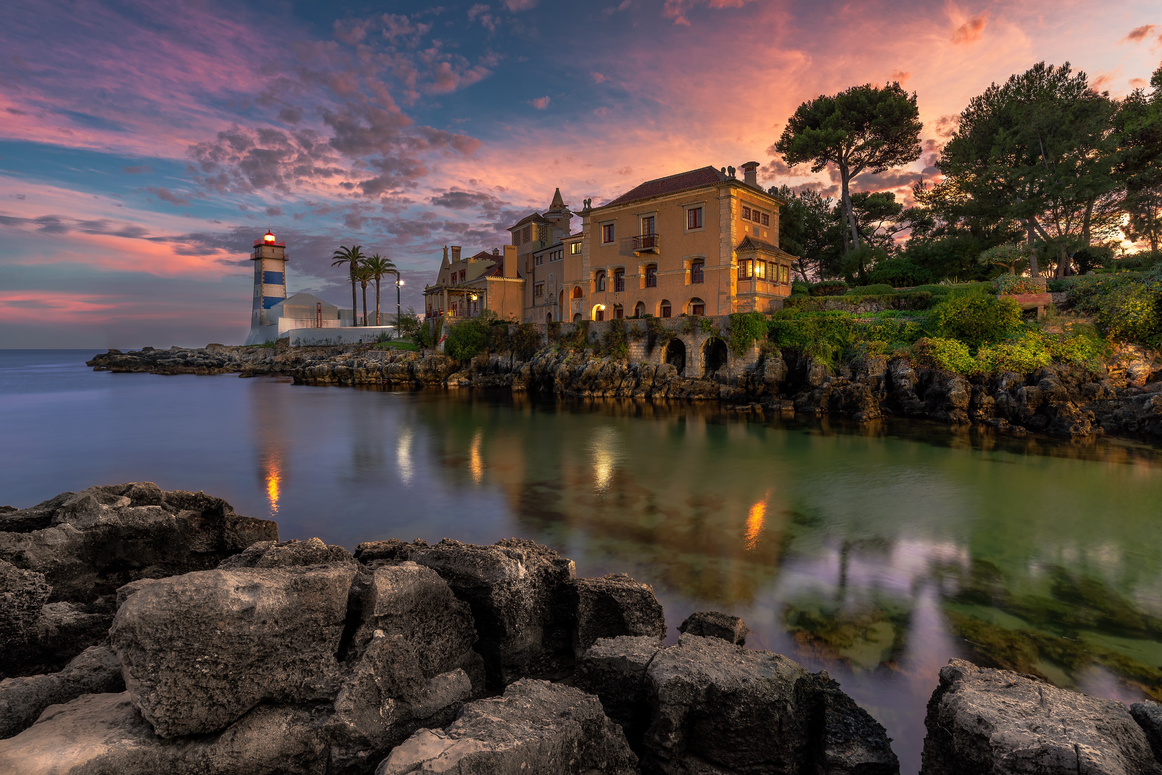 Lighthouse and houses on rocky coast at sunset in Cascais, Portugal HD Wallpaper