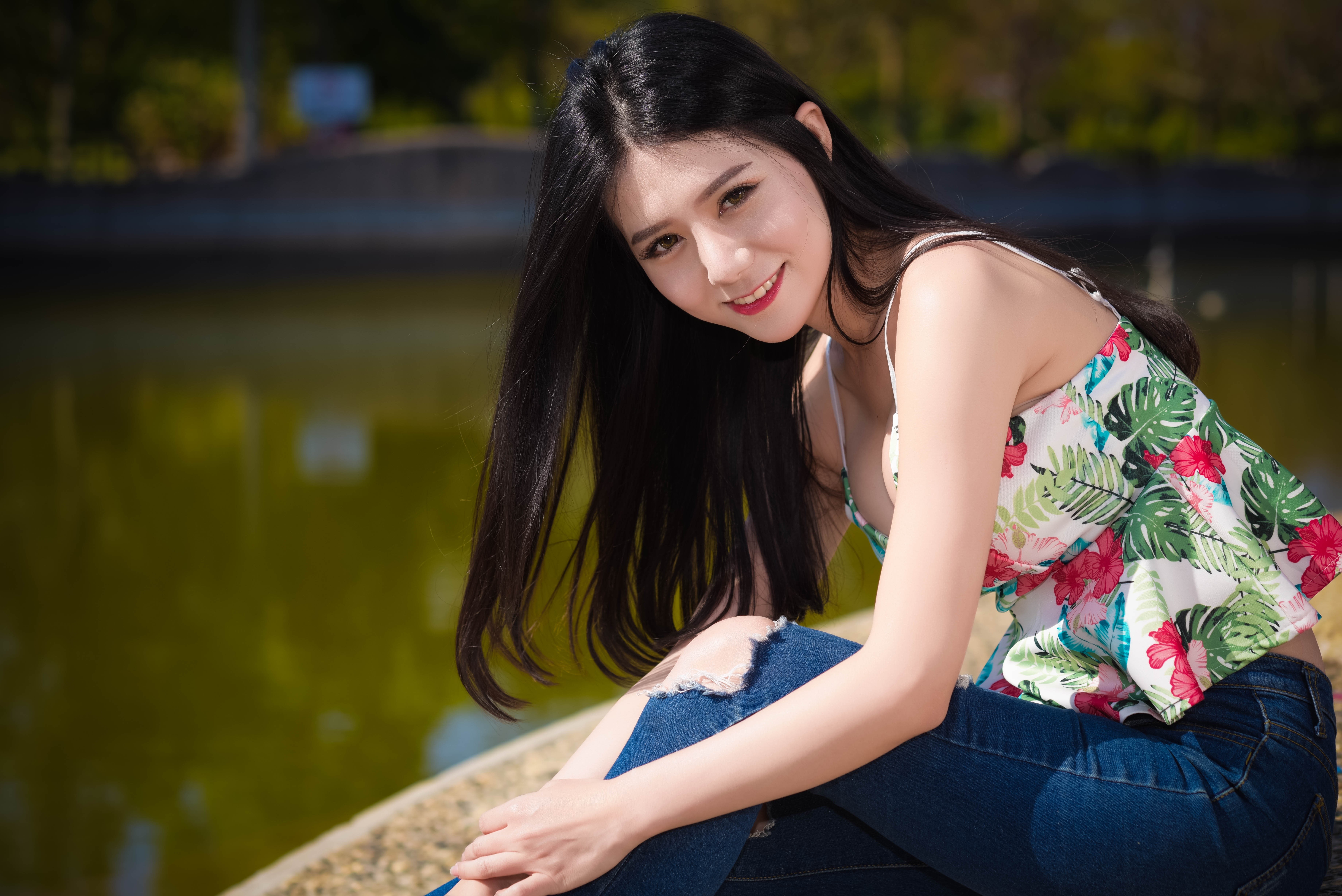 Brunette girl in jeans and floral top sitting by water HD Wallpaper