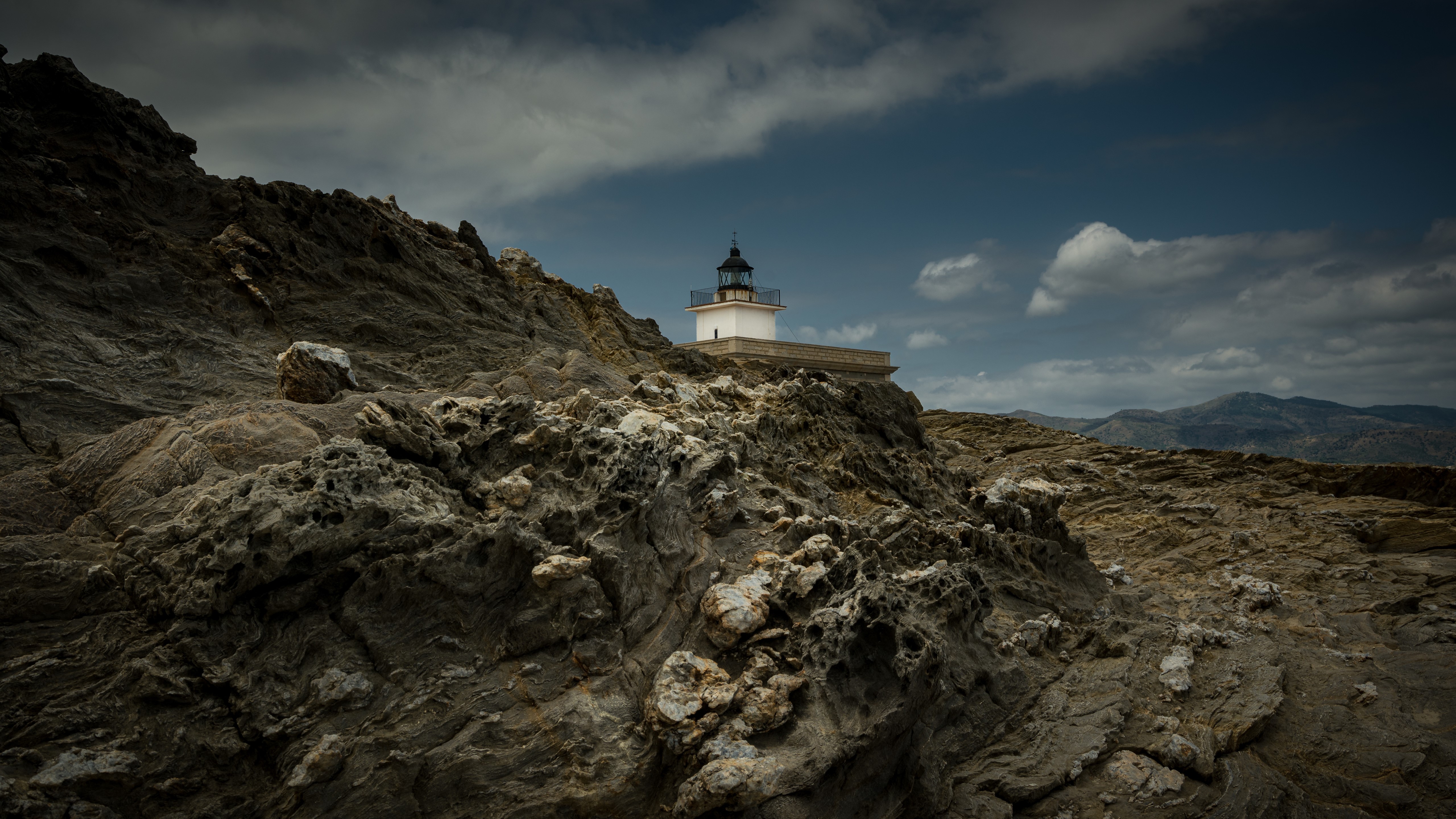 Lighthouse on a rocky coast under cloudy sky HD Wallpaper