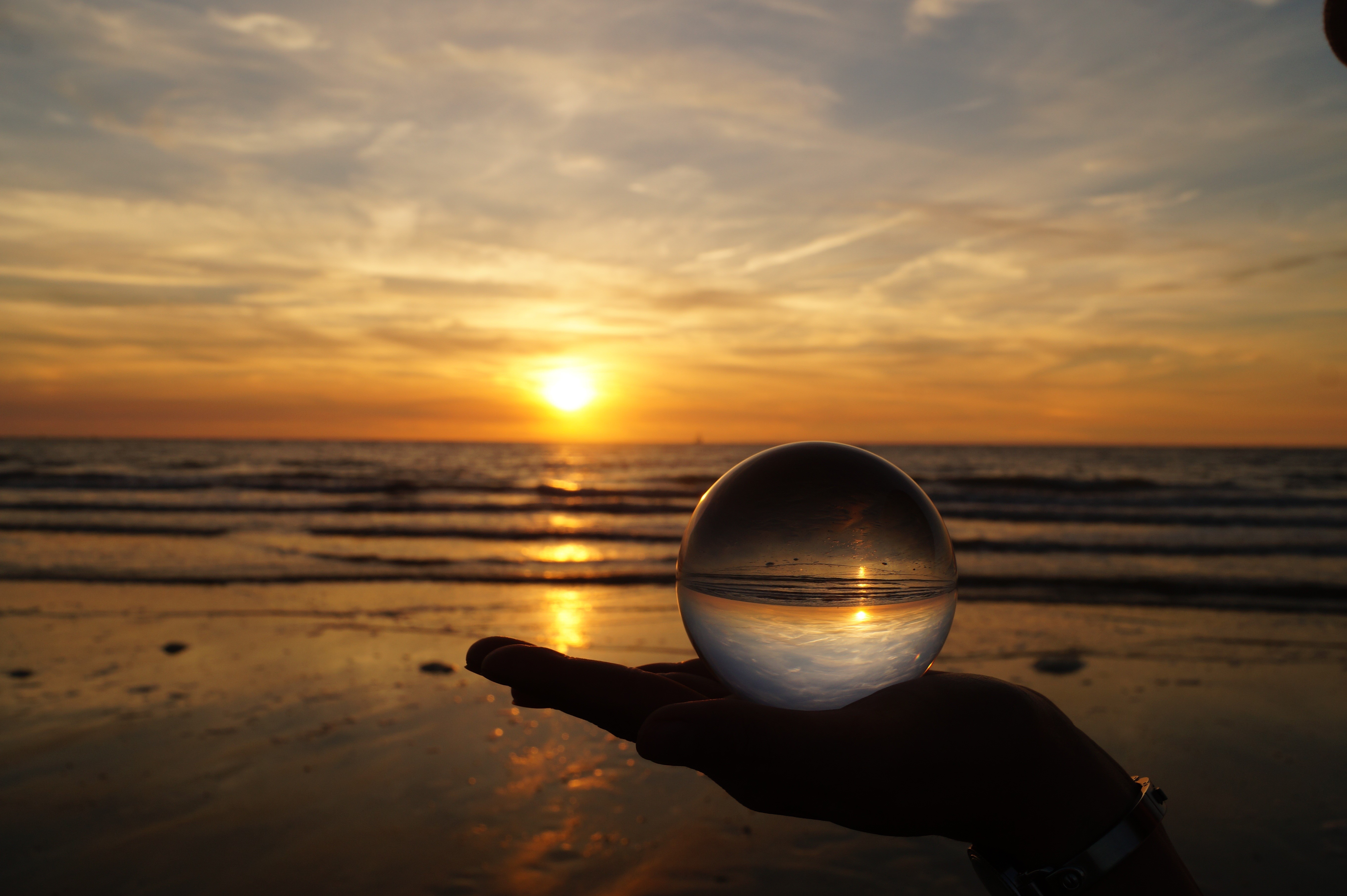Sunset over ocean reflected in glass ball held in hand HD Wallpaper