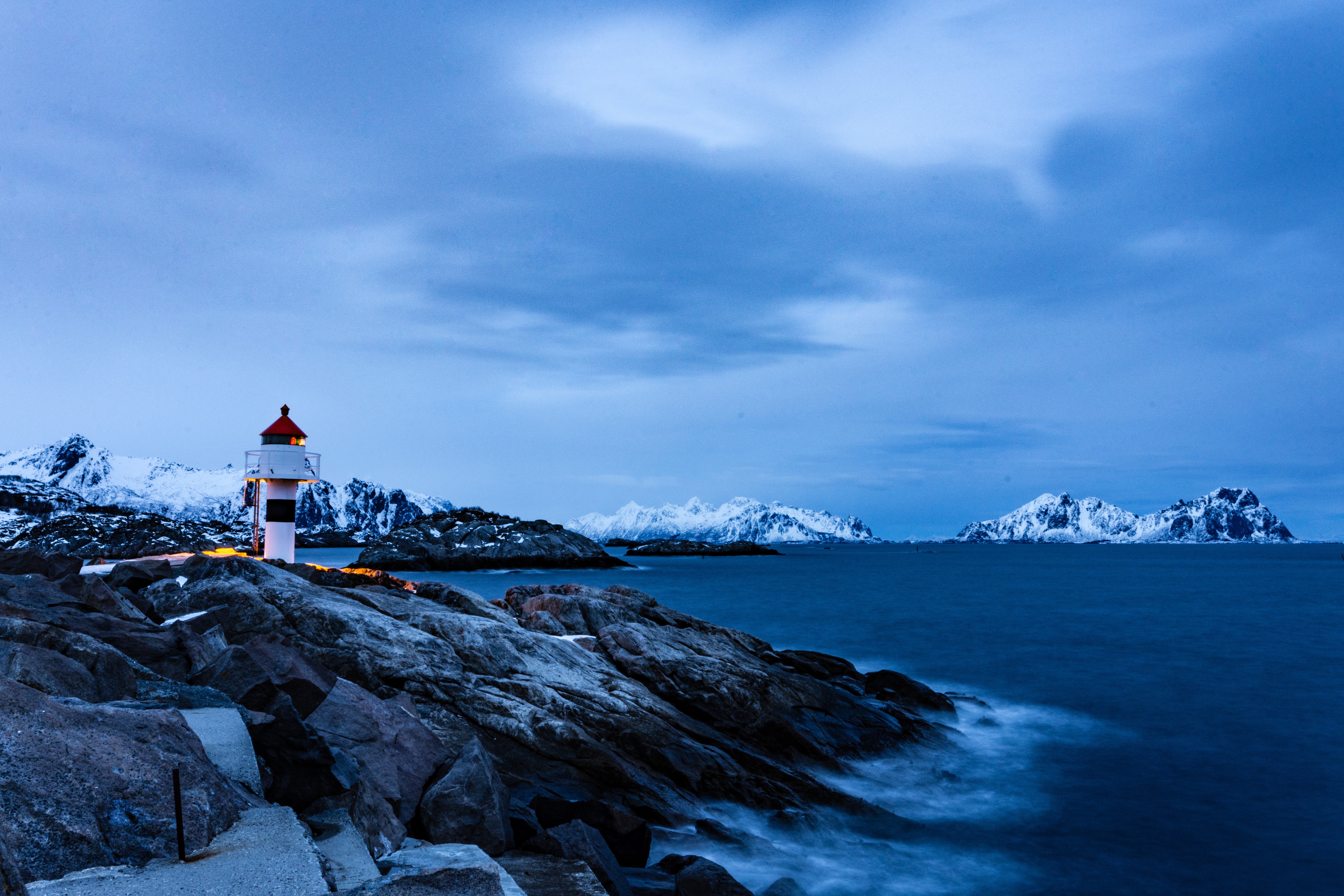 Lighthouse on rocky coast with snowy mountains in Norway HD Wallpaper