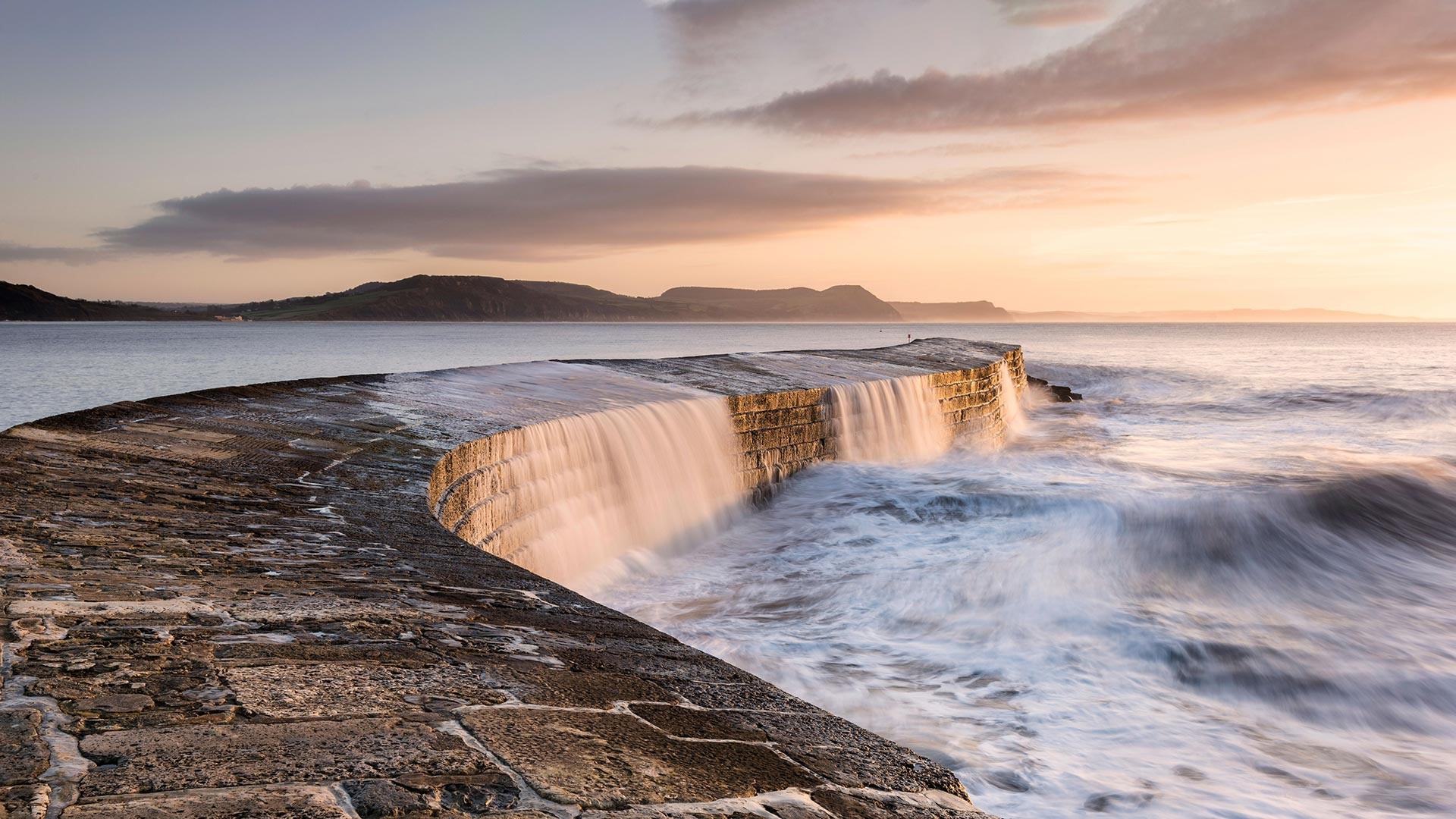 Curved stone pier with waves crashing at sunset HD Wallpaper