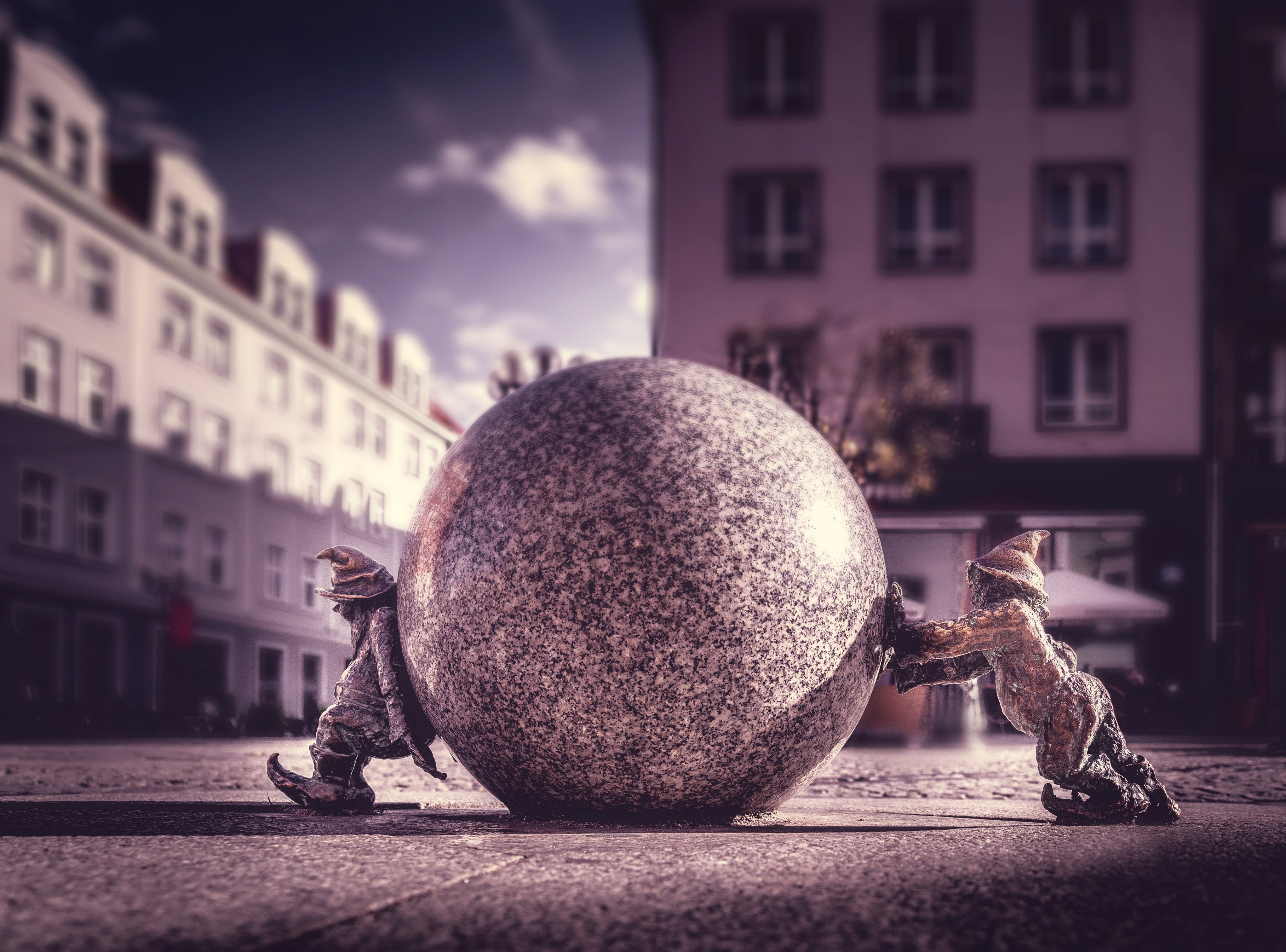 Two dwarf sculptures pushing a large stone ball in a city HD Wallpaper