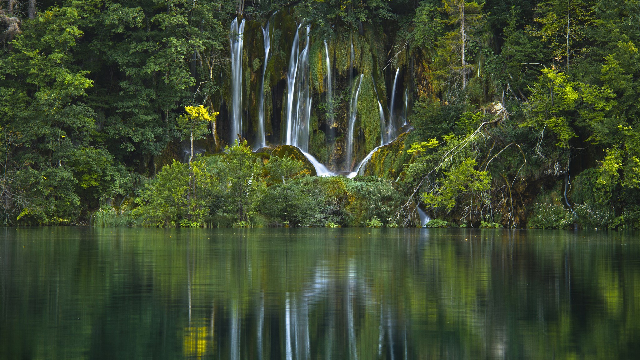 Waterfalls cascading into a serene lake surrounded by lush greenery HD Wallpaper