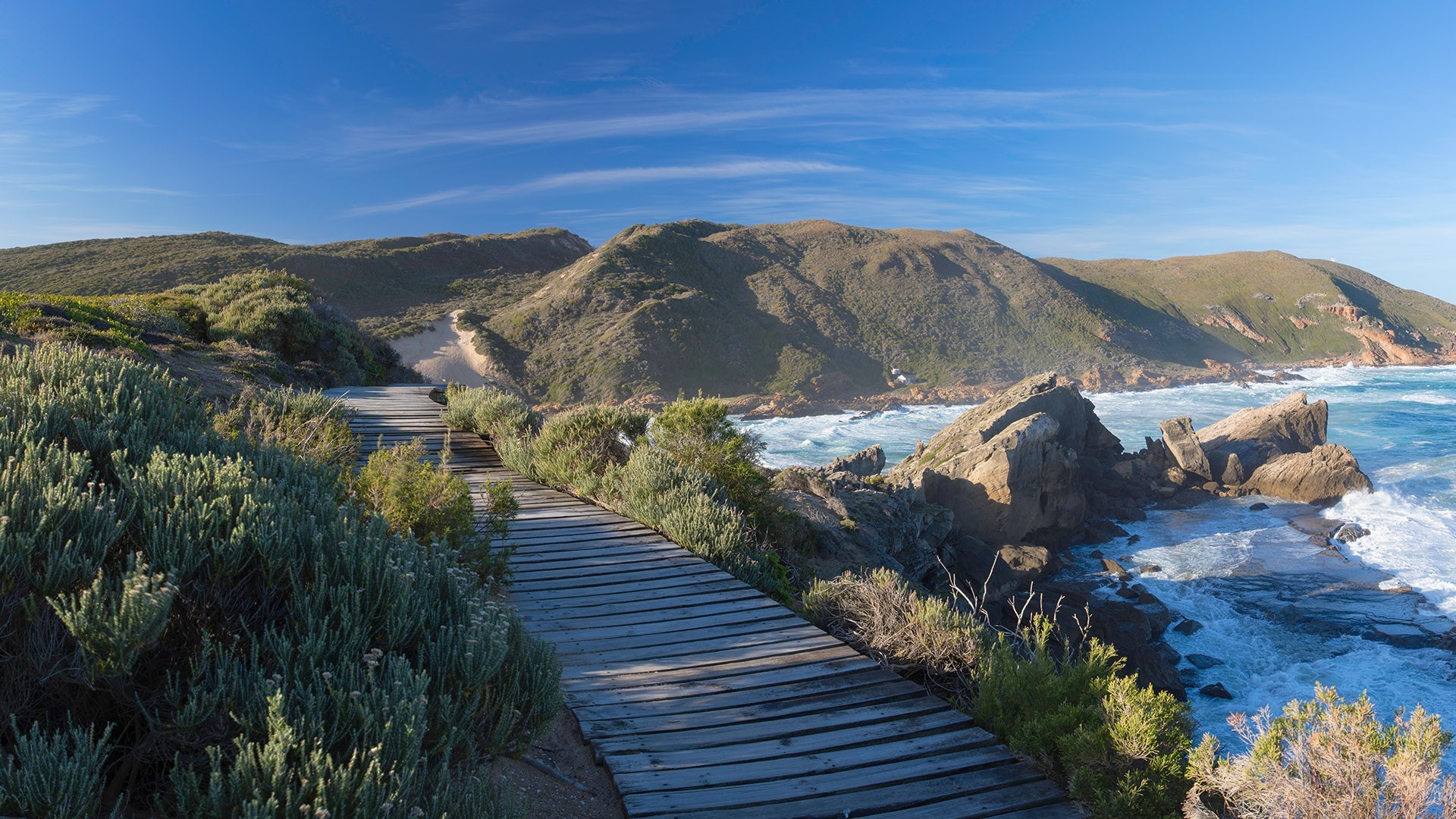 Wooden boardwalk along a rocky coastline with green hills HD Wallpaper