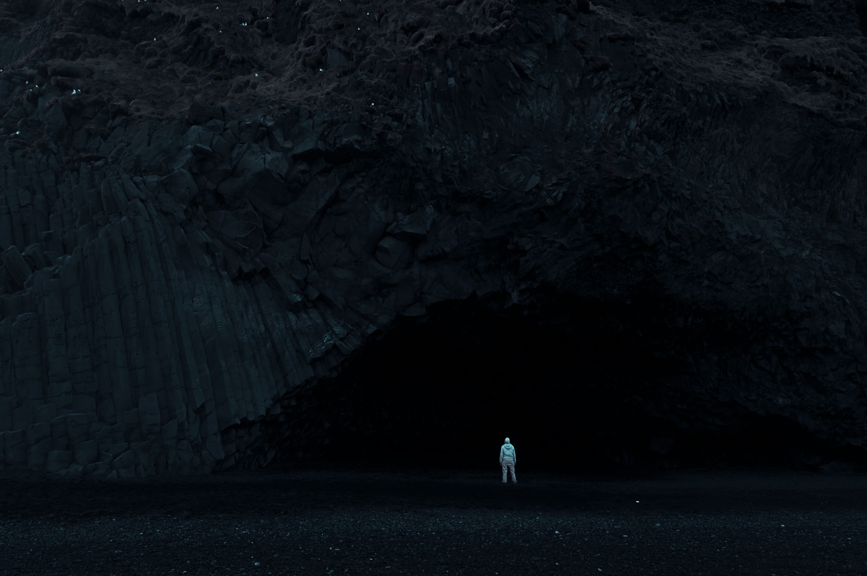 Person in a hazmat suit standing in front of a dark cave entrance HD Wallpaper