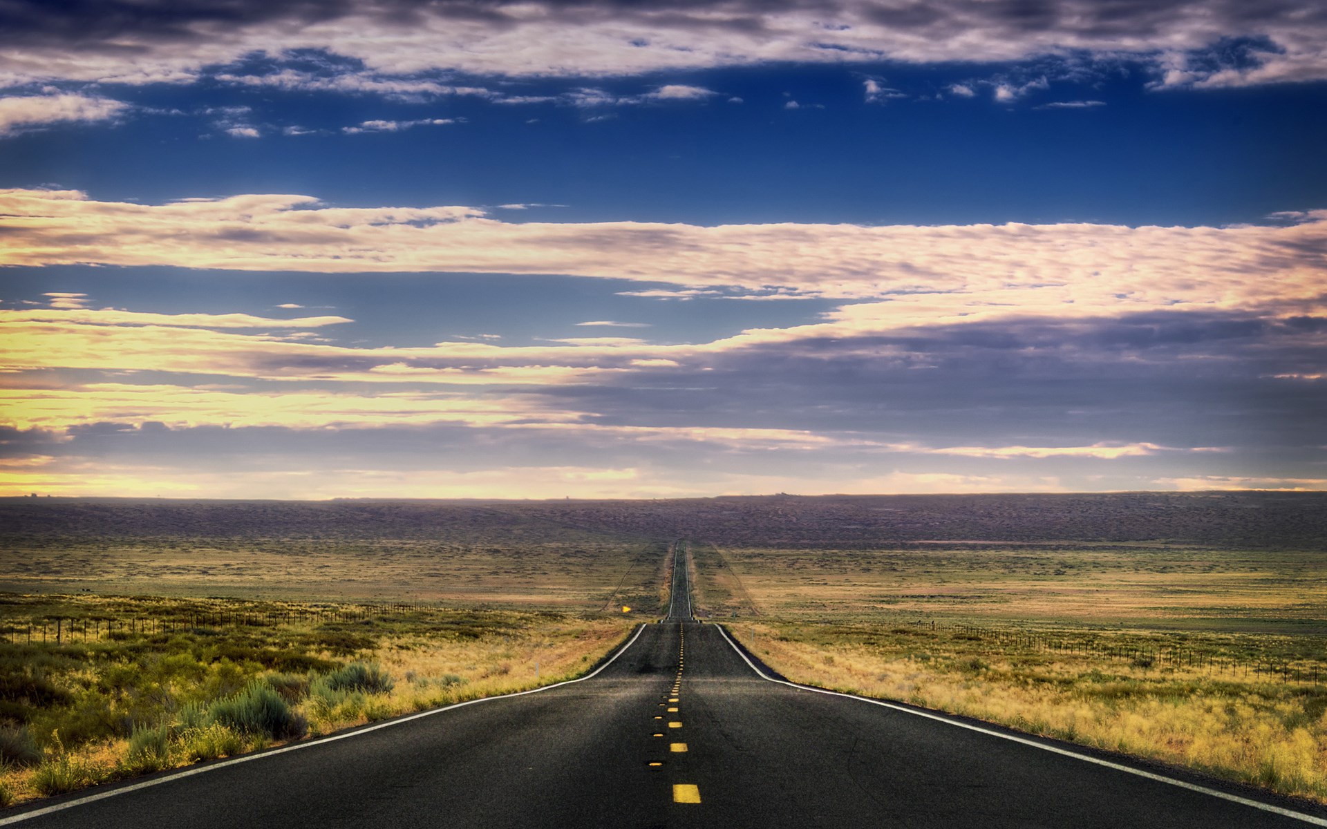 Endless road through a vast, grassy plain under a cloudy sky HD Wallpaper