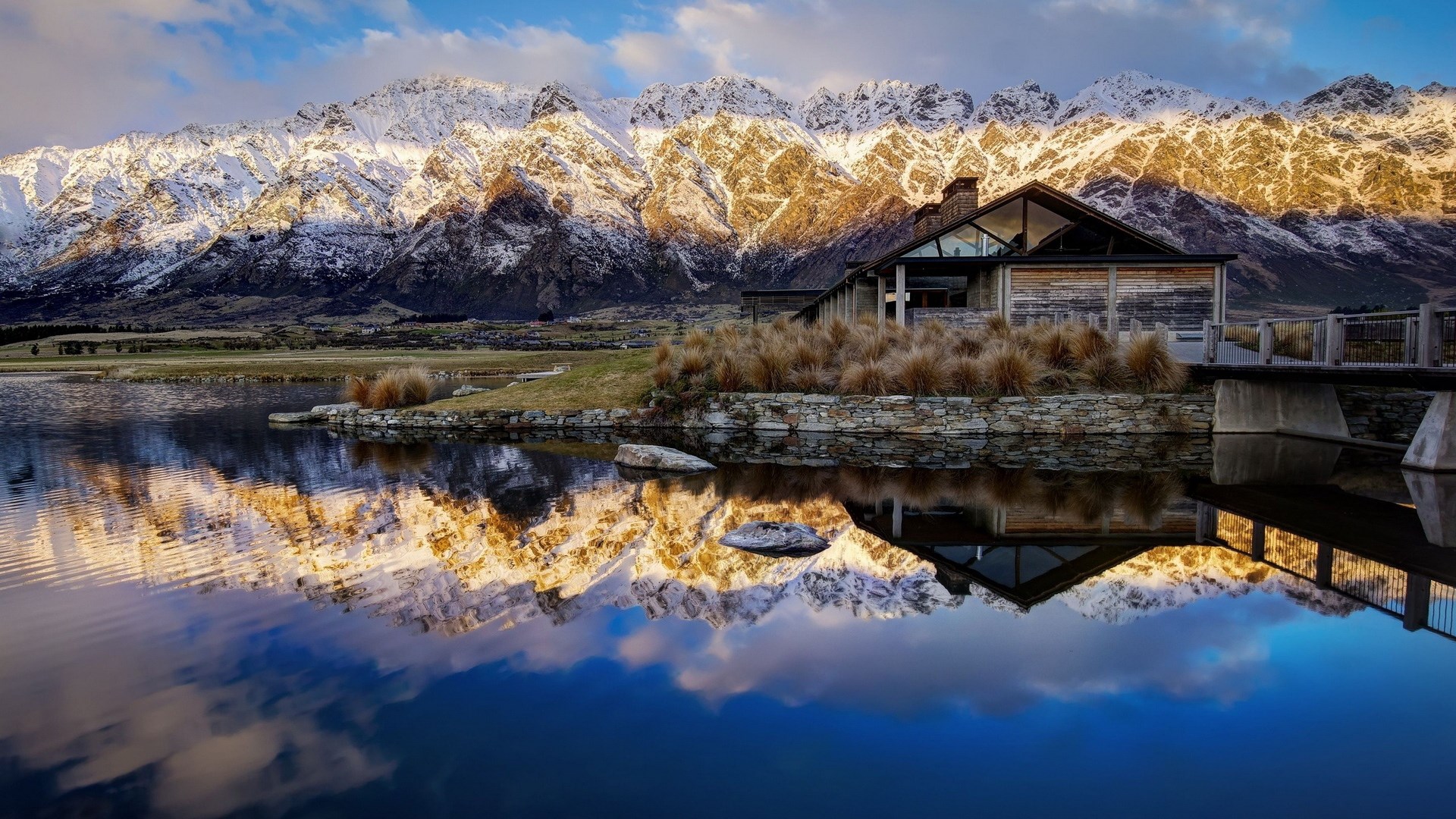 Modern building by a lake with snow-capped mountains HD Wallpaper