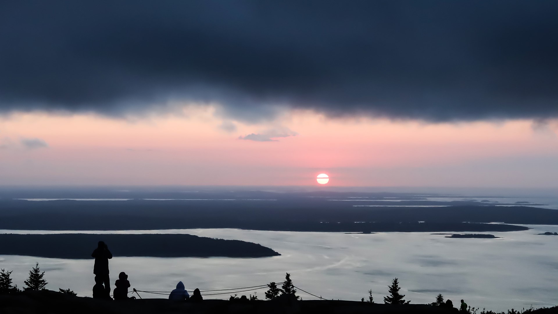 People silhouetted on a hill watching a sunset over water HD Wallpaper
