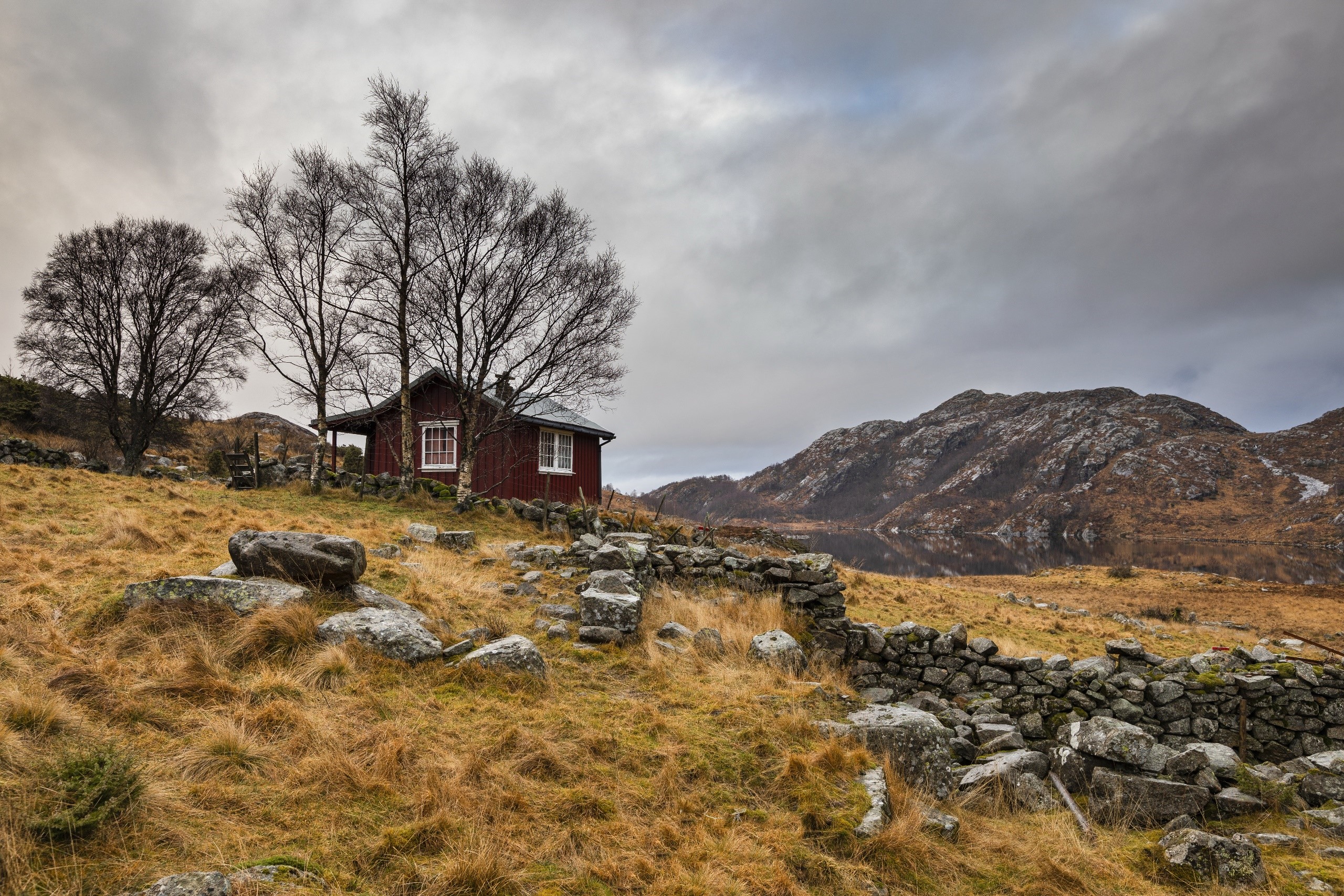 Red cabin in a rocky, grassy landscape with mountains and a lake HD Wallpaper
