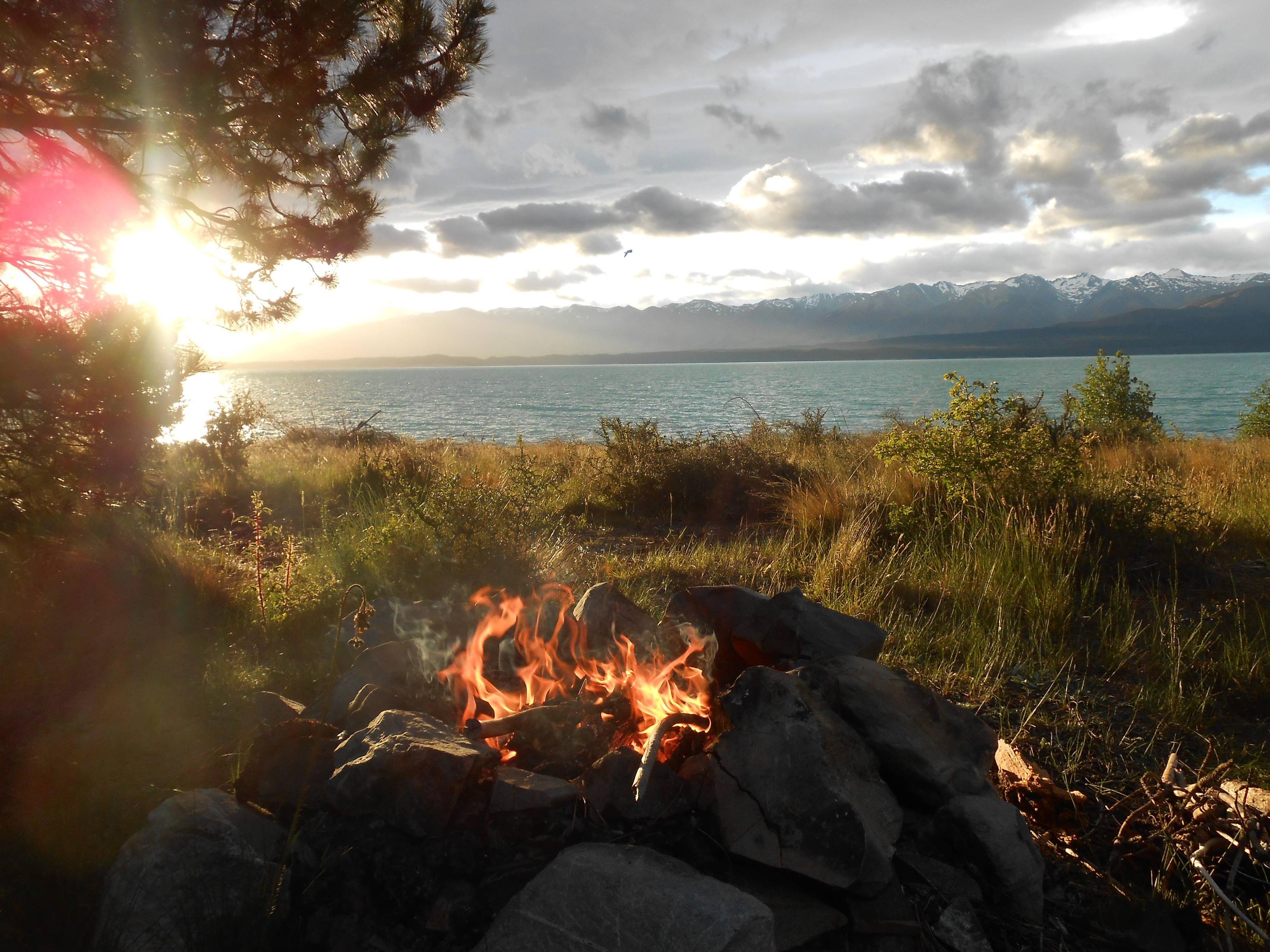 Campfire by a lake with mountains at sunset HD Wallpaper