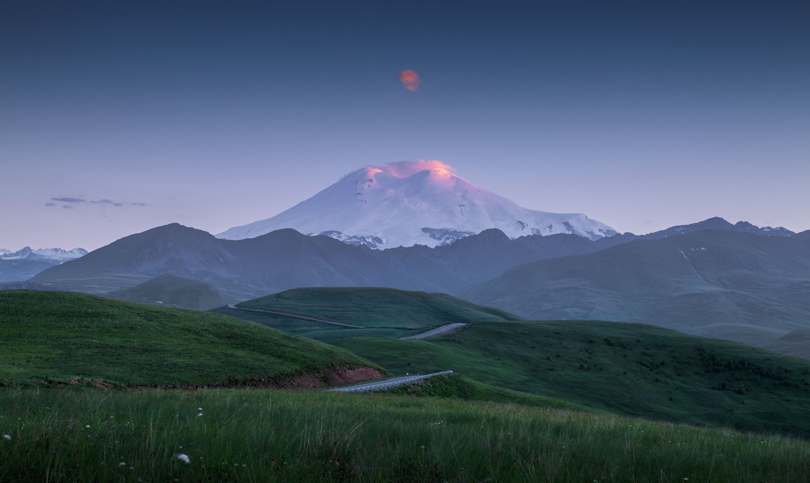 Mount Elbrus at sunrise with rolling green hills and a road HD Wallpaper
