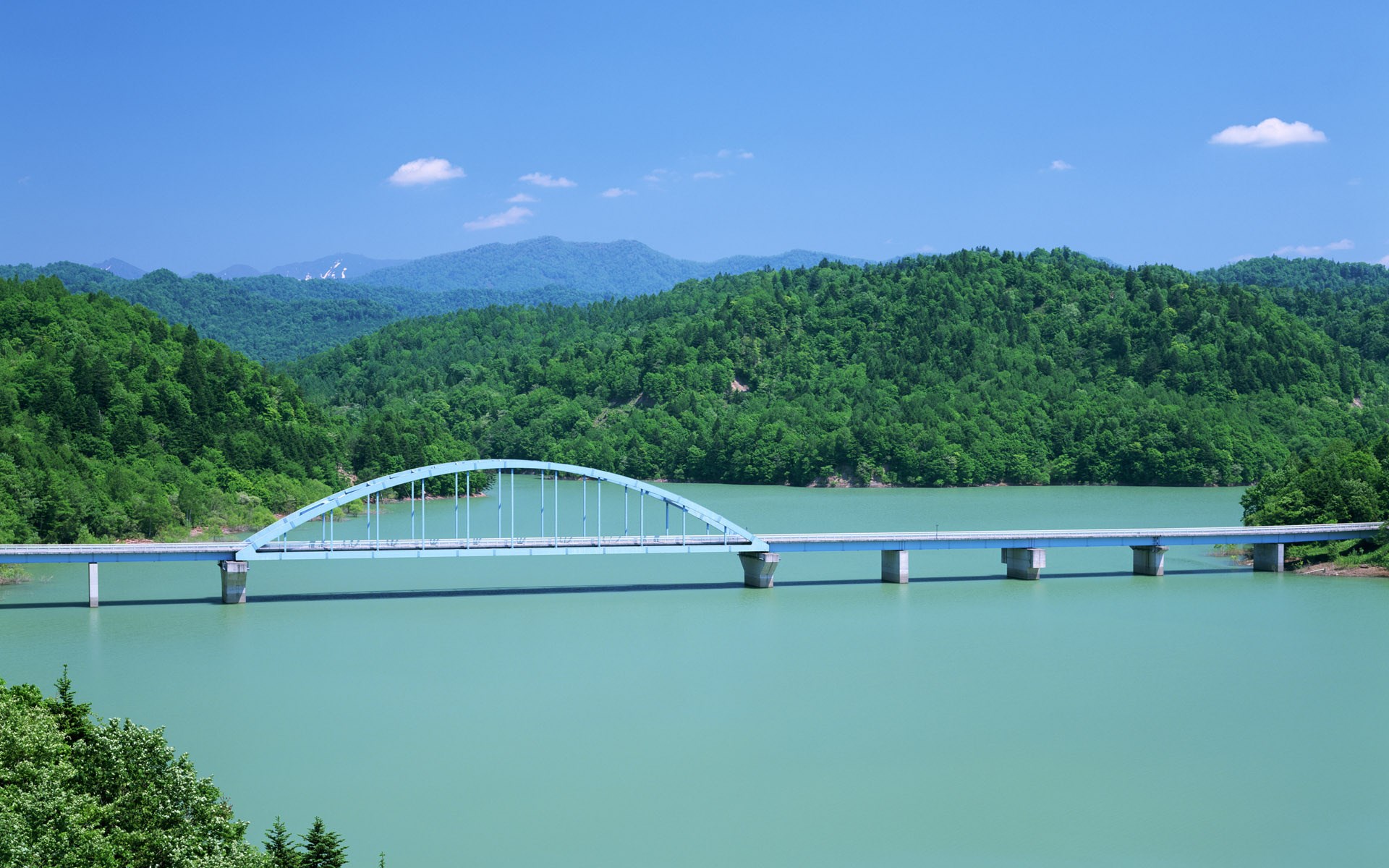 Arch bridge over a turquoise lake with forested mountains HD Wallpaper