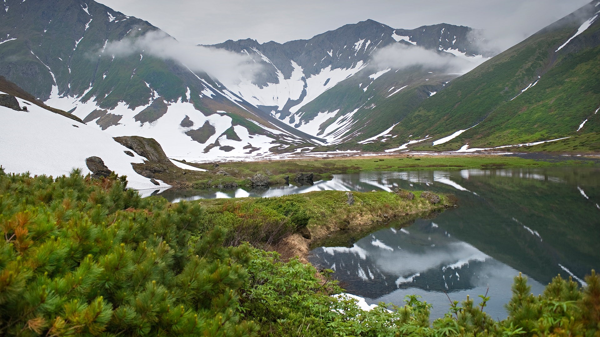 Snow-capped mountains reflected in a calm lake surrounded by greenery HD Wallpaper