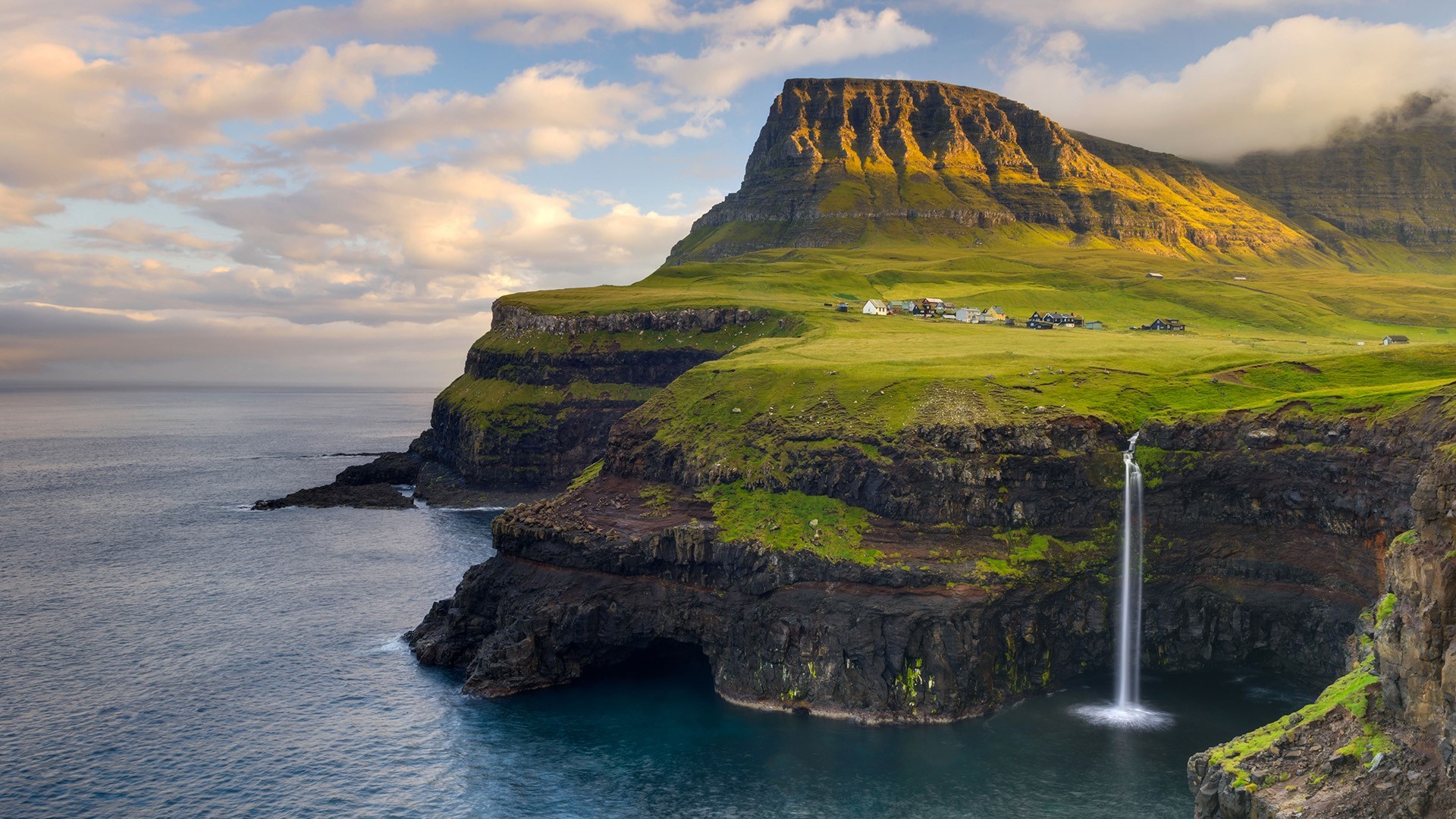 Waterfall cascading into the sea near a village in the Faroe Islands HD Wallpaper