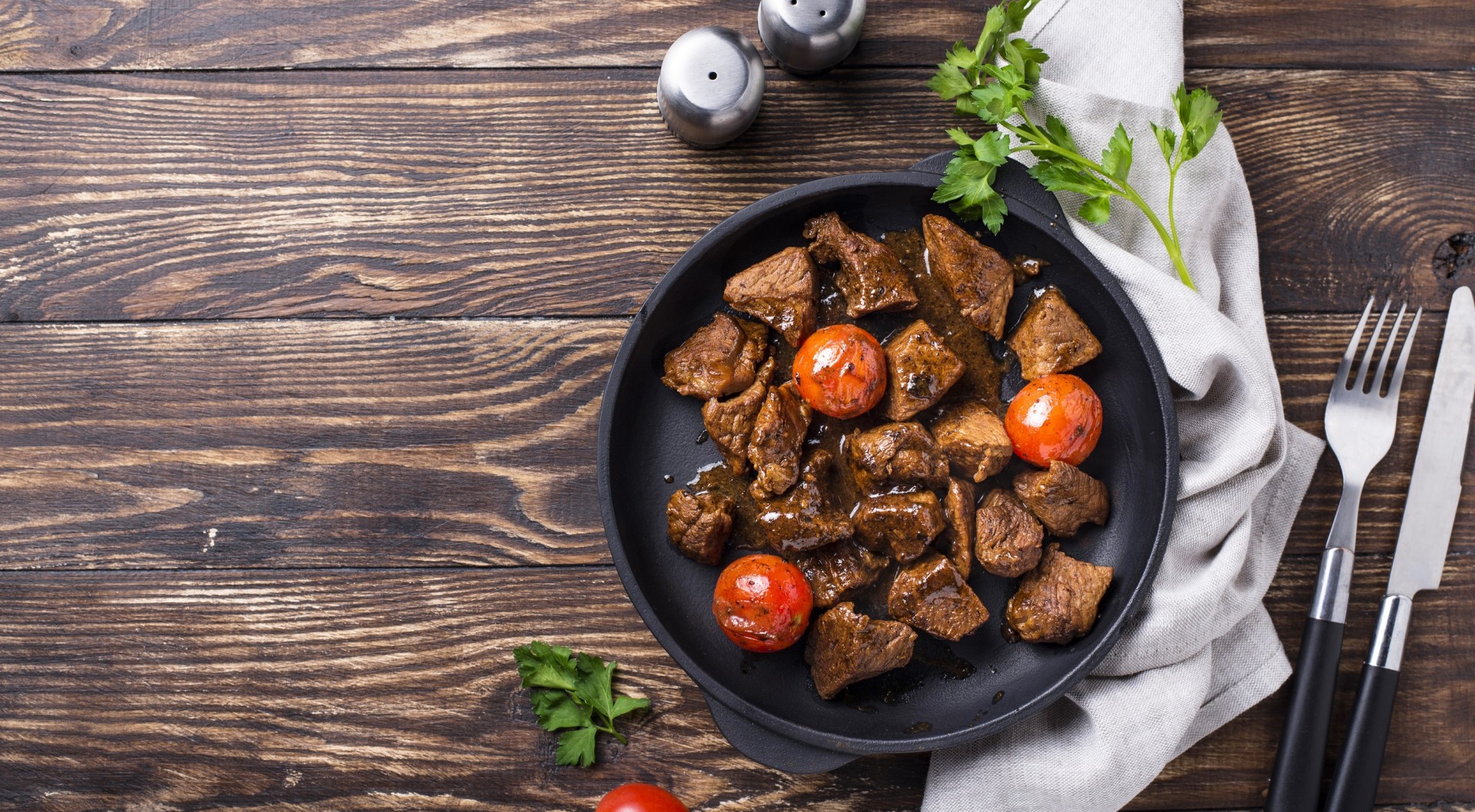 Meat and tomatoes in a pan with cutlery on a wooden table HD Wallpaper