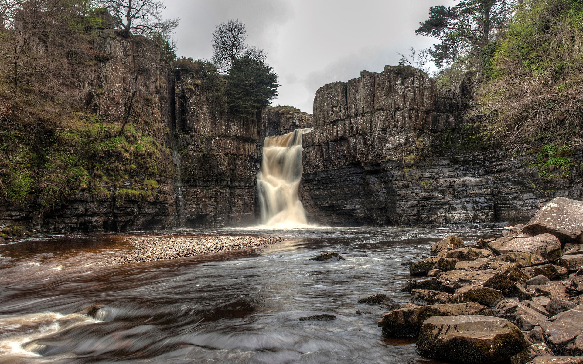 Waterfall cascading over rocky cliffs into a river HD Wallpaper