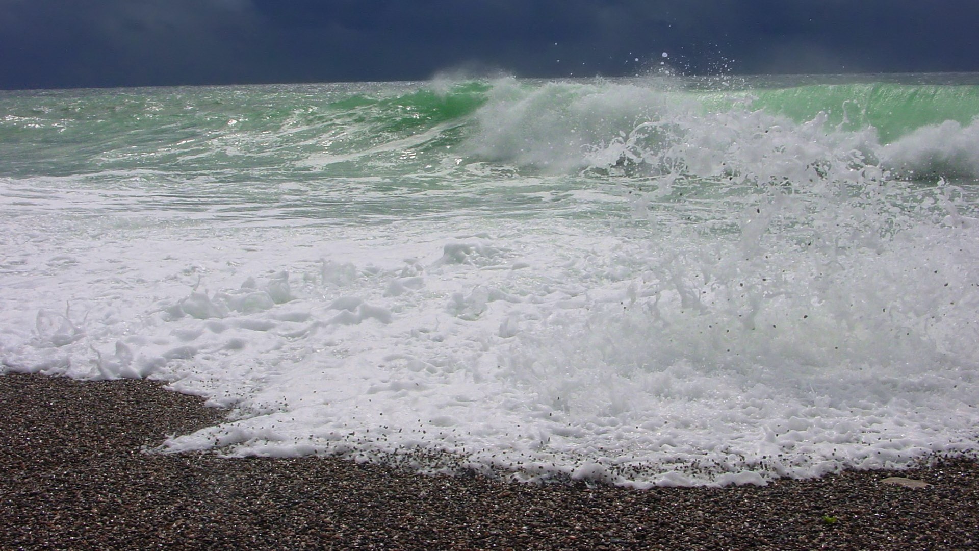 Waves crashing on a pebble beach under a dark sky HD Wallpaper