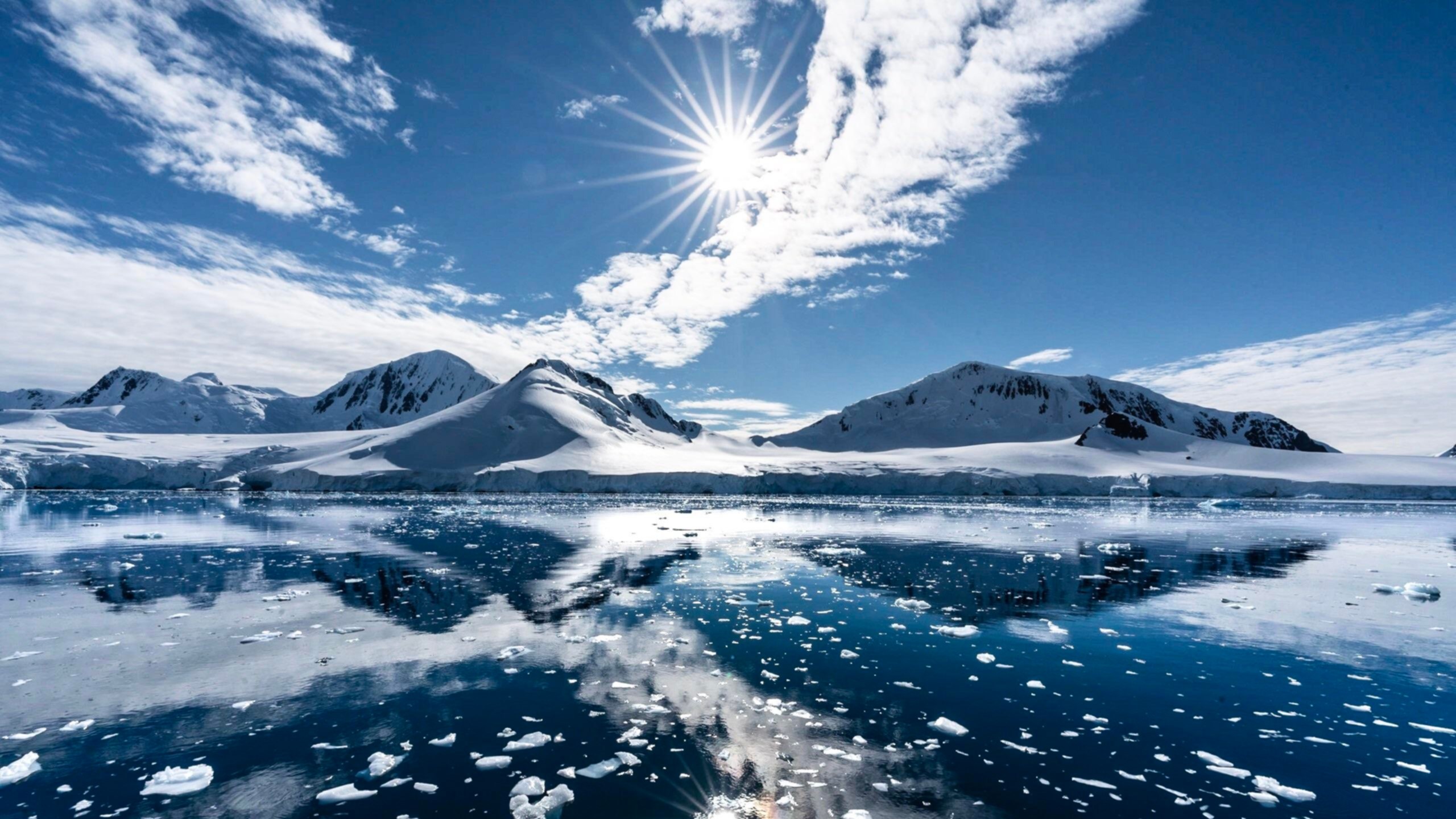 Snowy mountains reflected in icy water under a sunny sky HD Wallpaper