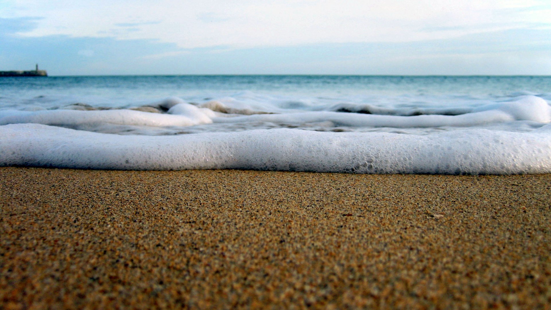 Ocean waves washing onto a sandy beach with a lighthouse in the distance HD Wallpaper