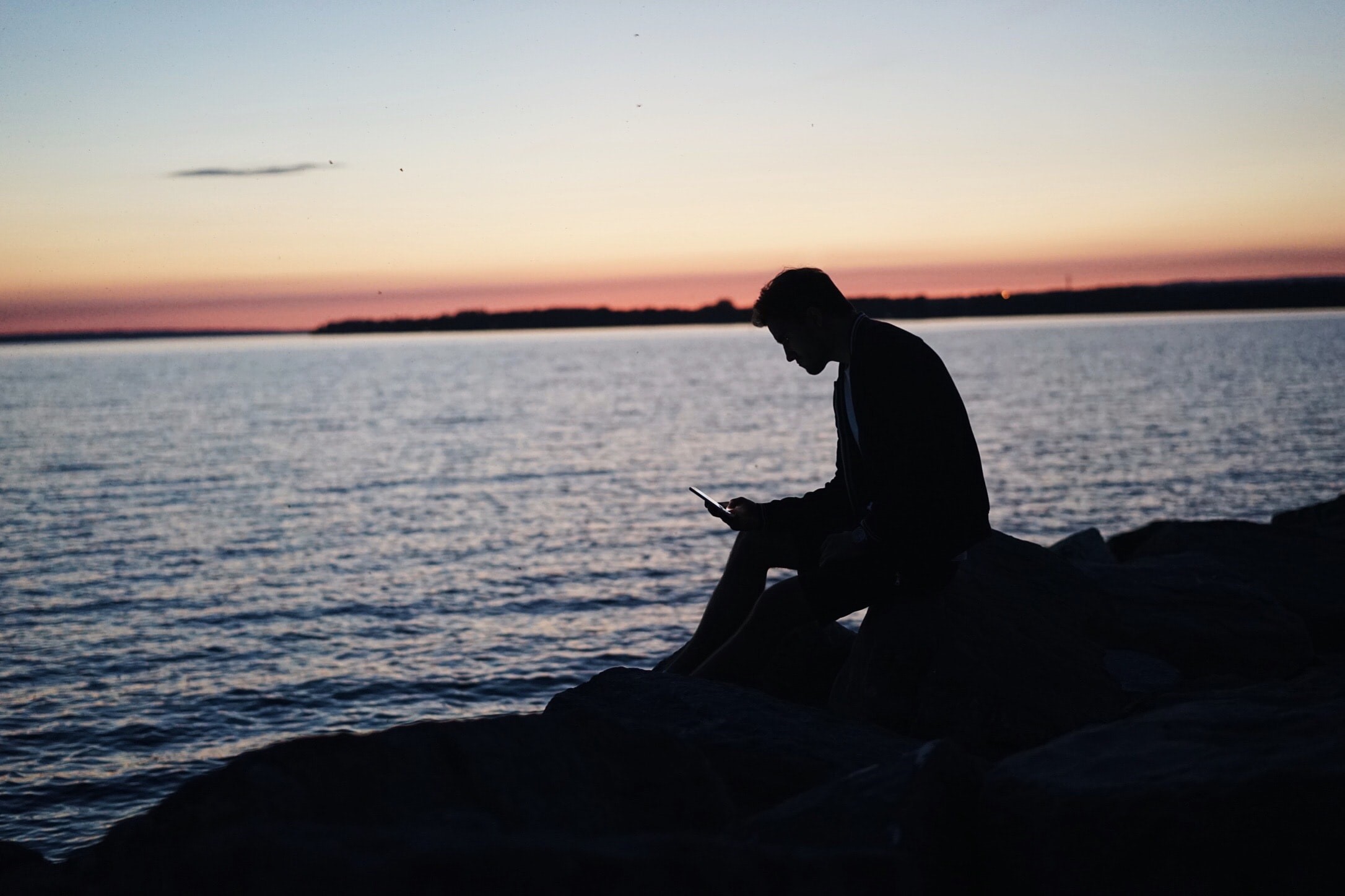 Silhouette of a man on rocks using a phone at sunset HD Wallpaper