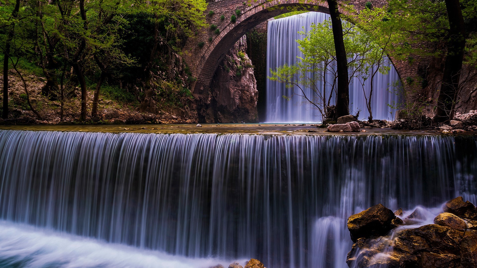 Waterfall flowing under a stone bridge in a forest HD Wallpaper