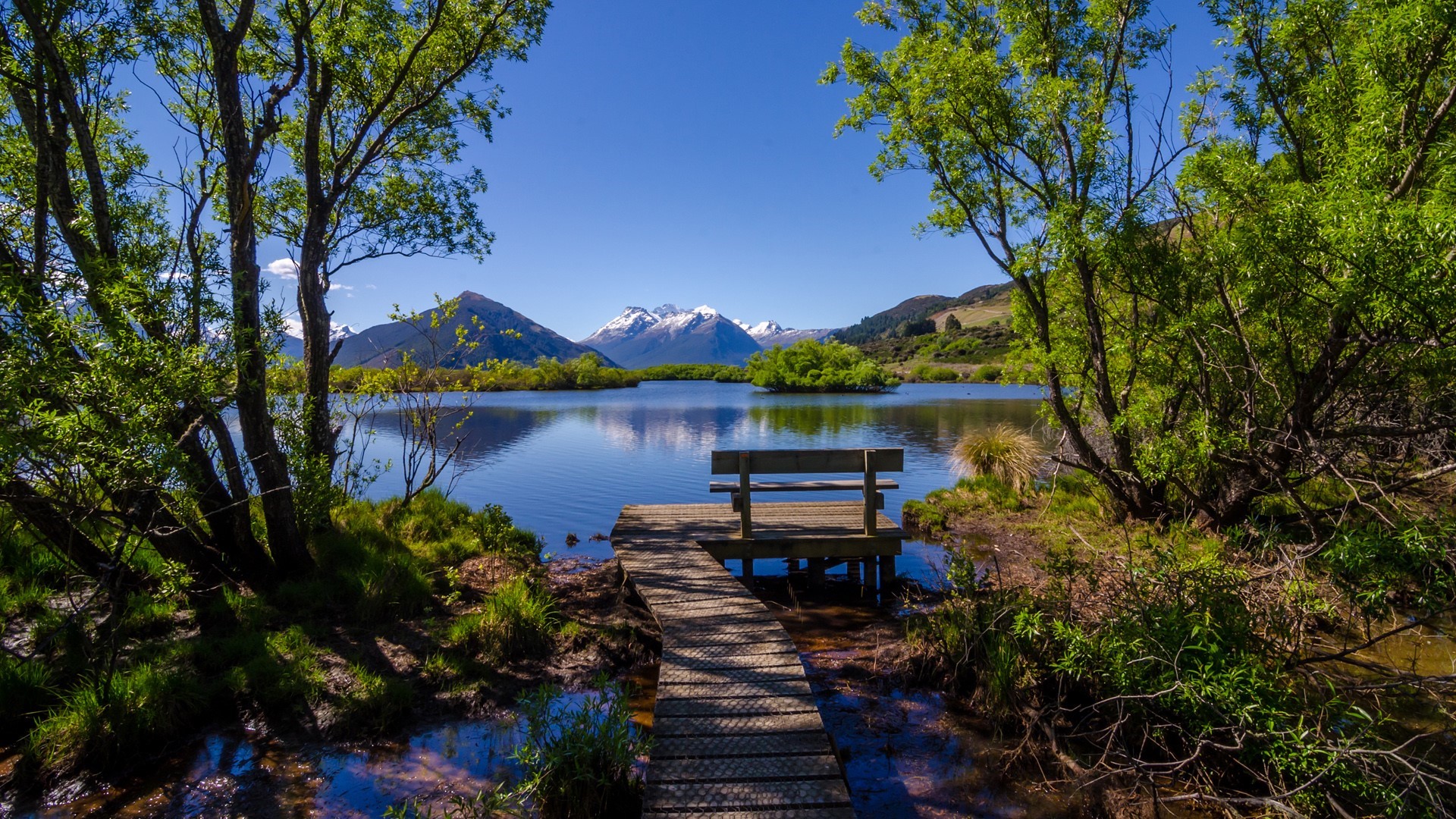 Wooden walkway and bench by a lake with snowy mountains HD Wallpaper