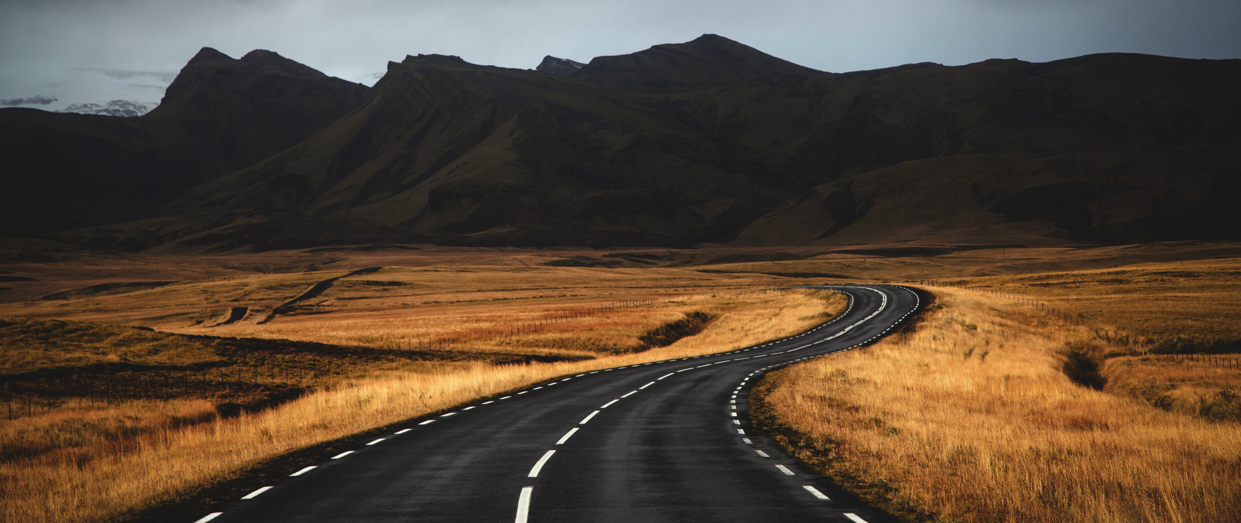 Winding road through a golden field towards dark mountains HD Wallpaper